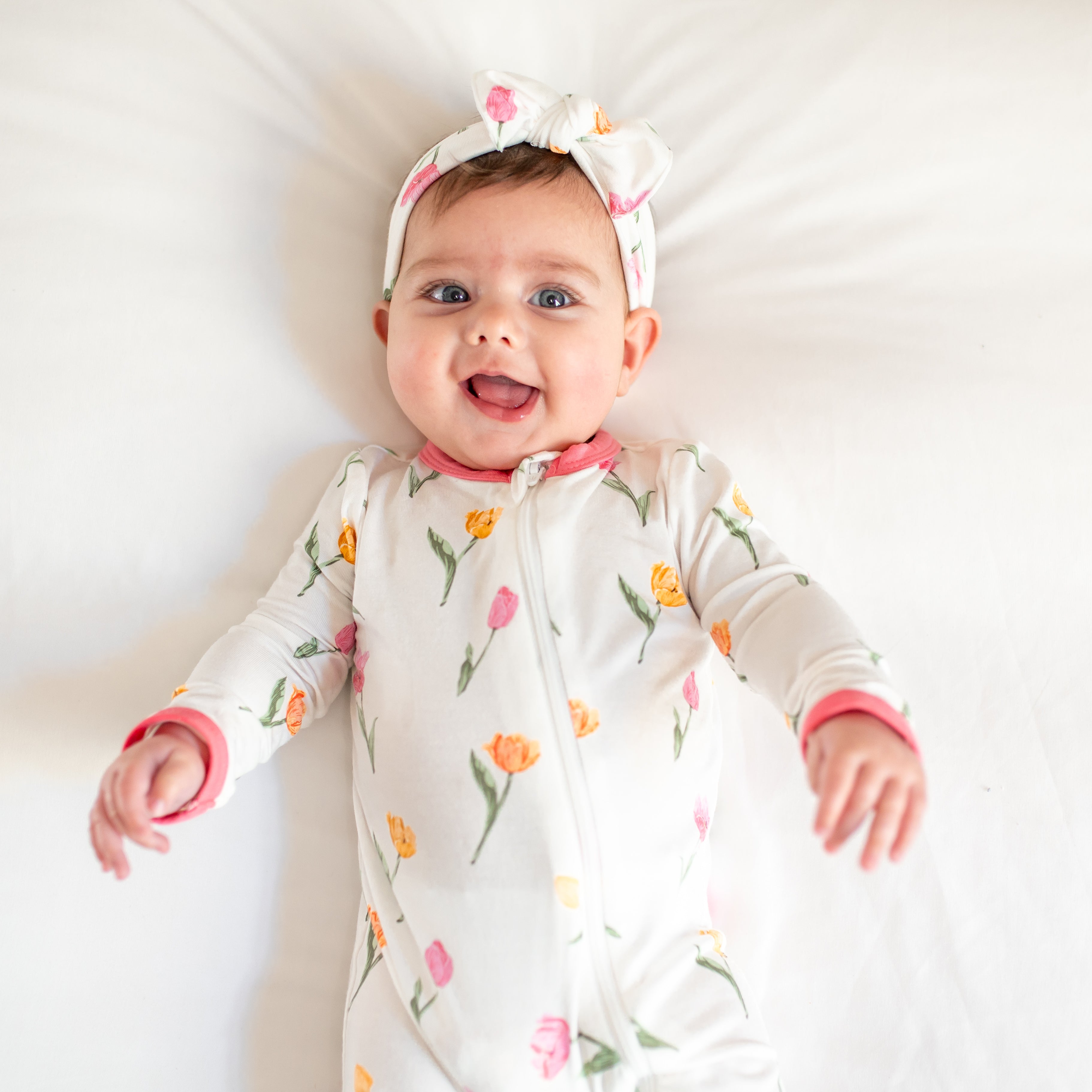 Close up of a toddler laying on a bed wearing the Zippered Romper in Tulip with matching knotted bow headband