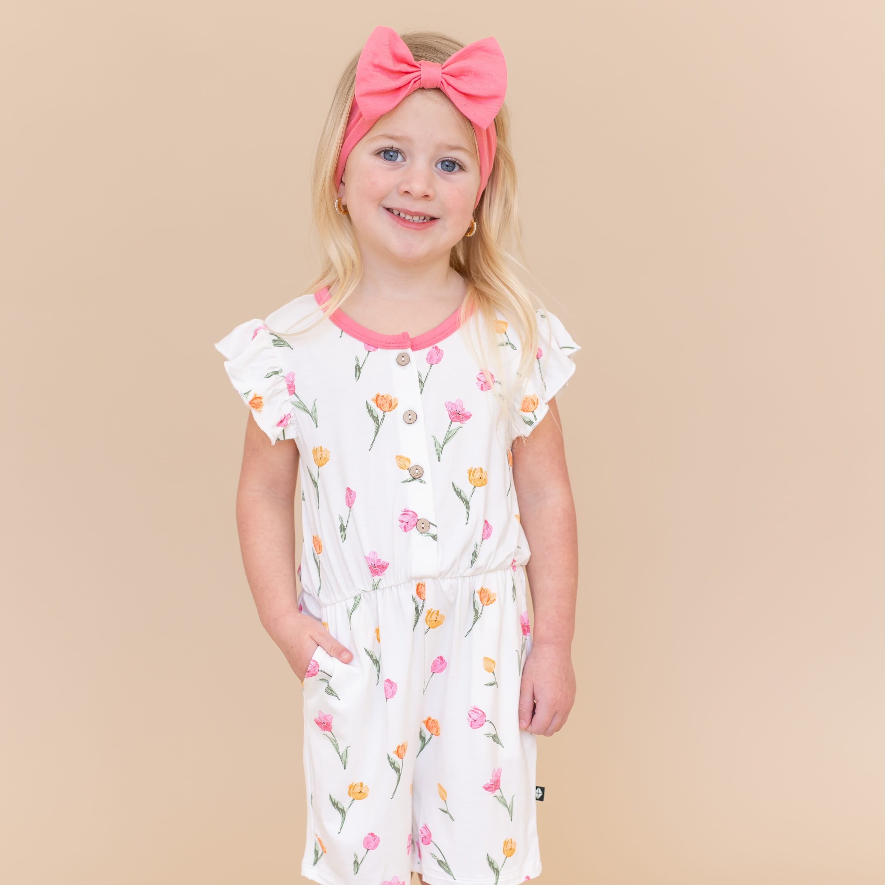 Young girl posing wearing the Flutter Sleeve Romper in Tulip paired with a pink bow