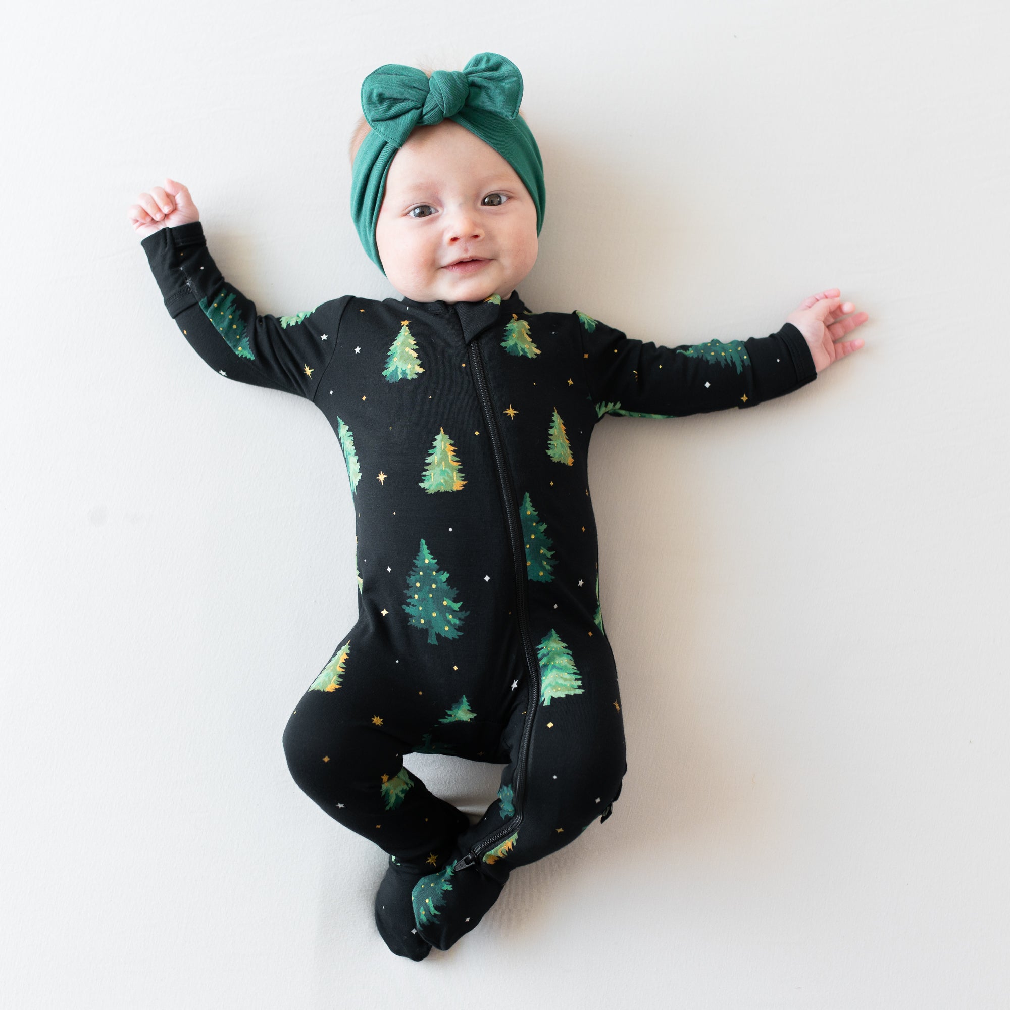 Infant laying down wearing the Zippered Footie in Twinkle Tree with Emerald knotted bow headband