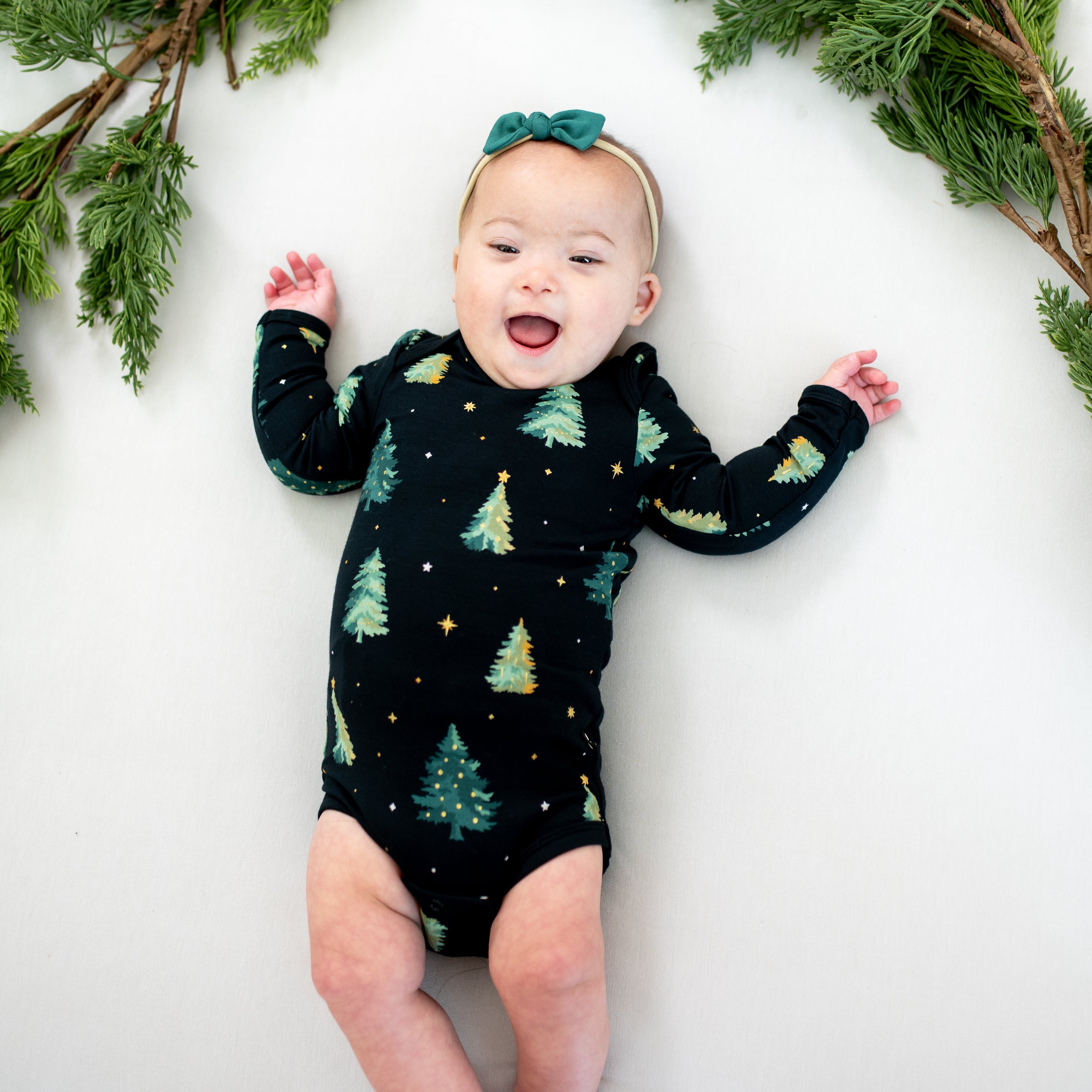 Smiling infant laying down with greenery around her head wearing the Long Sleeve Bodysuit in Twinkle Tree and Tiny Nylon bow in Emerald