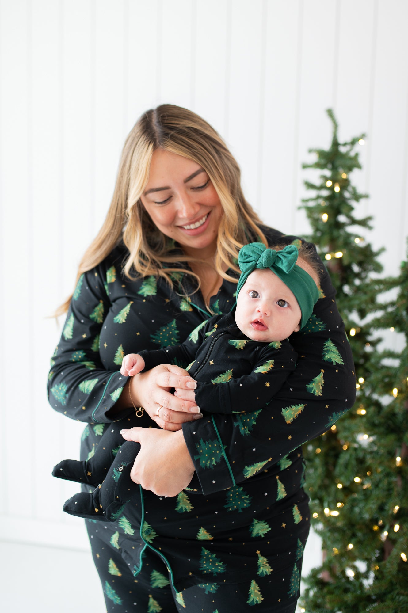 Mother wearing the Long-Sleeved Women's Pajama Set in Twinkle Tree holding her infant daughter who is wearing a matching footie and green bow