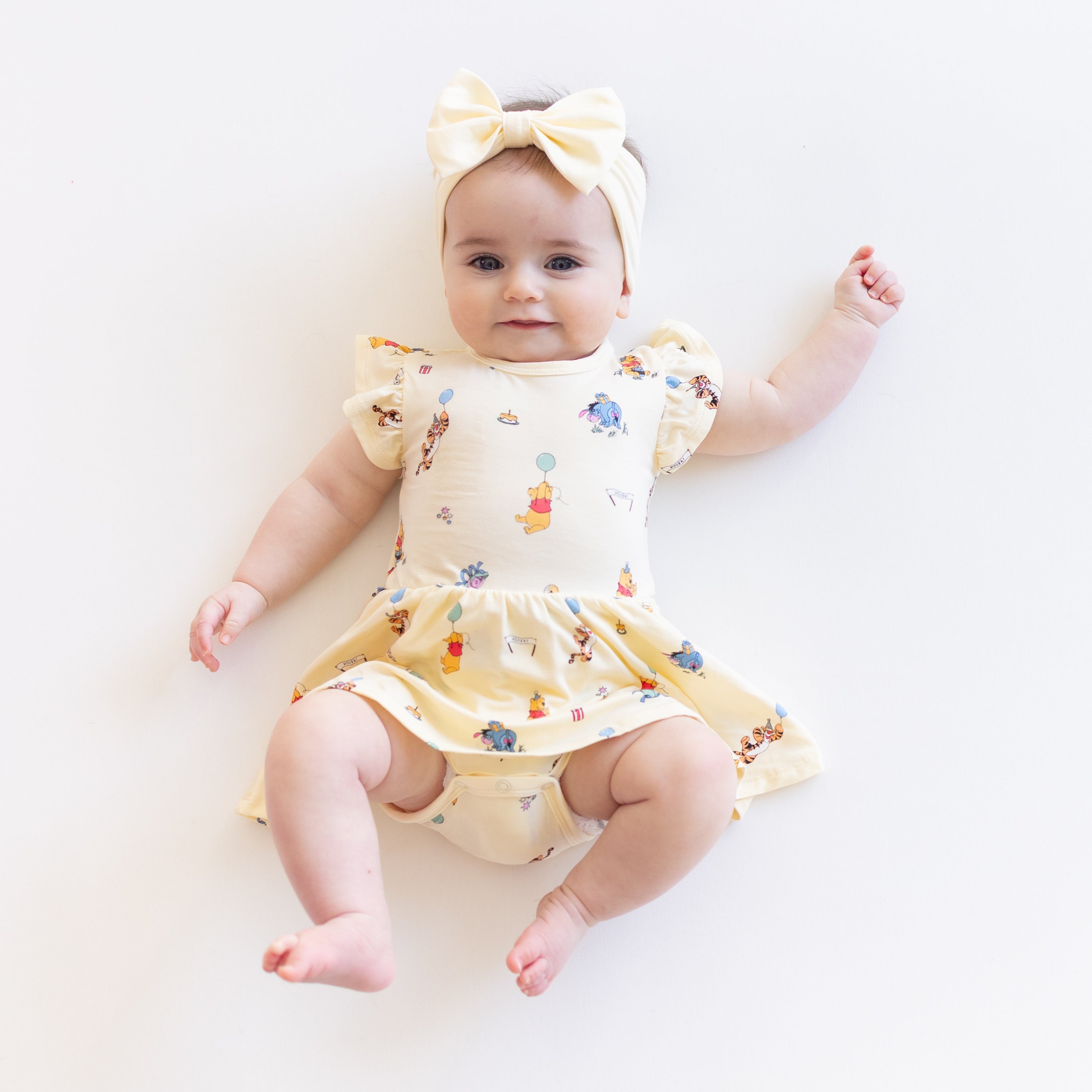 Smiling infant laying on a white surface wearing the Twirl Bodysuit Dress in Winnie the Pooh Celebration with a light yellow bow