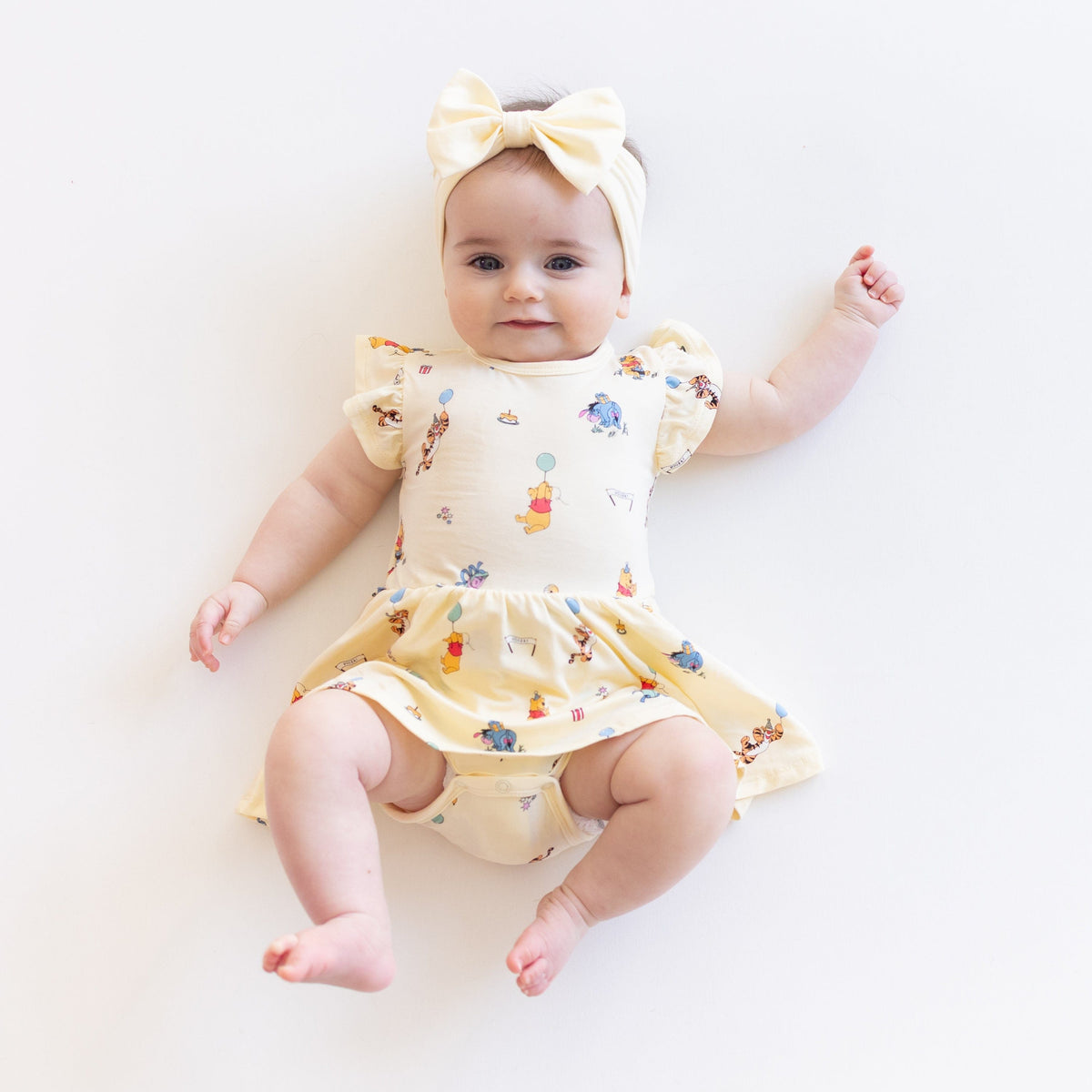 Smiling infant laying on a white surface wearing the Twirl Bodysuit Dress in Winnie the Pooh Celebration with a light yellow bow