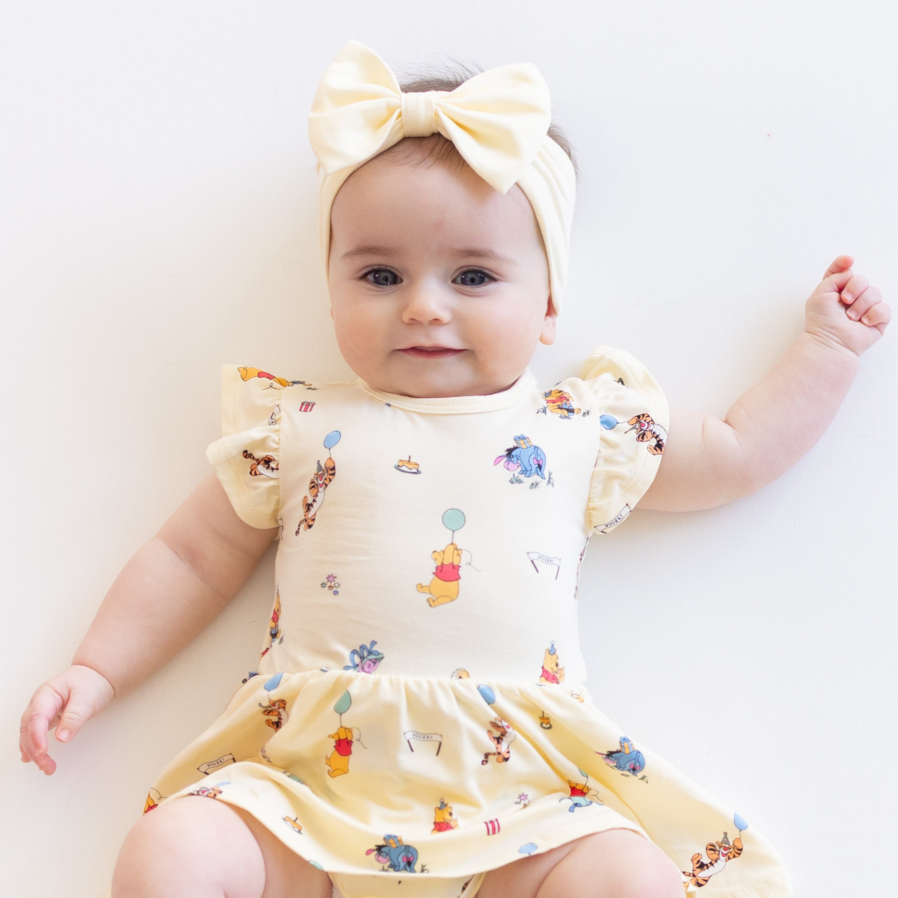 Close up of infant wearing the Twirl Bodysuit Dress in Winnie the Pooh Celebration with a light yellow bow