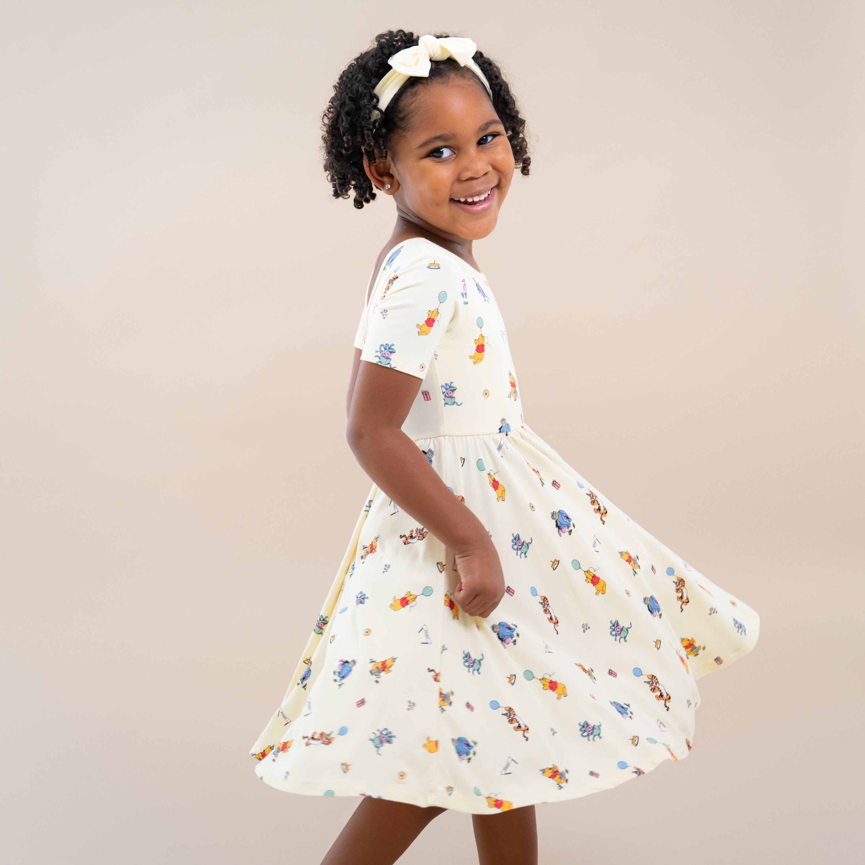 Smiling girl standing in front of a light neutral surface wearing the Twirl Dress in Winnie the Pooh Celebration