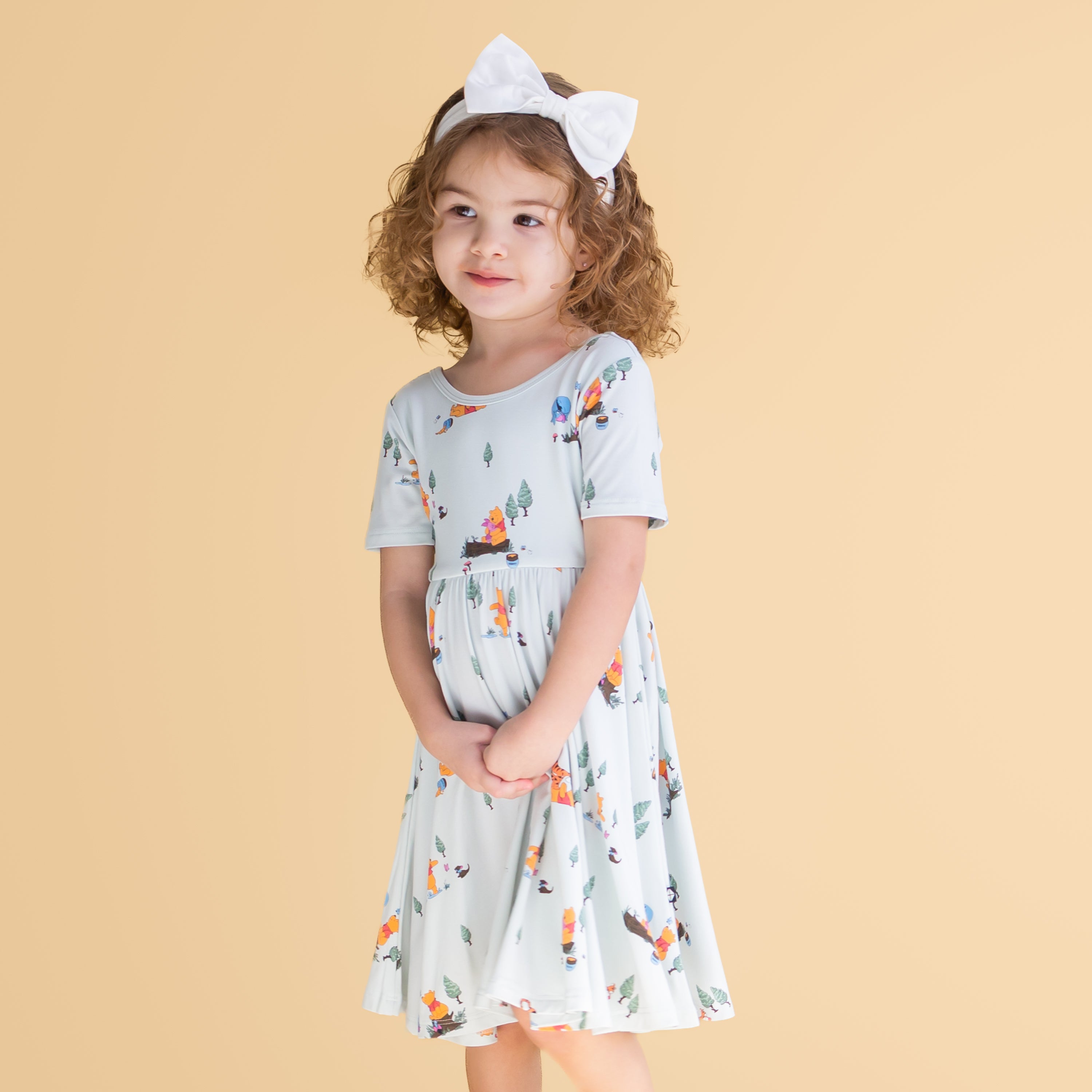 Young girl posing wearing the Twirl Dress in Winnie the Pooh Hundred Acre Wood paired with a white bow in front of a mustard yellow backgound