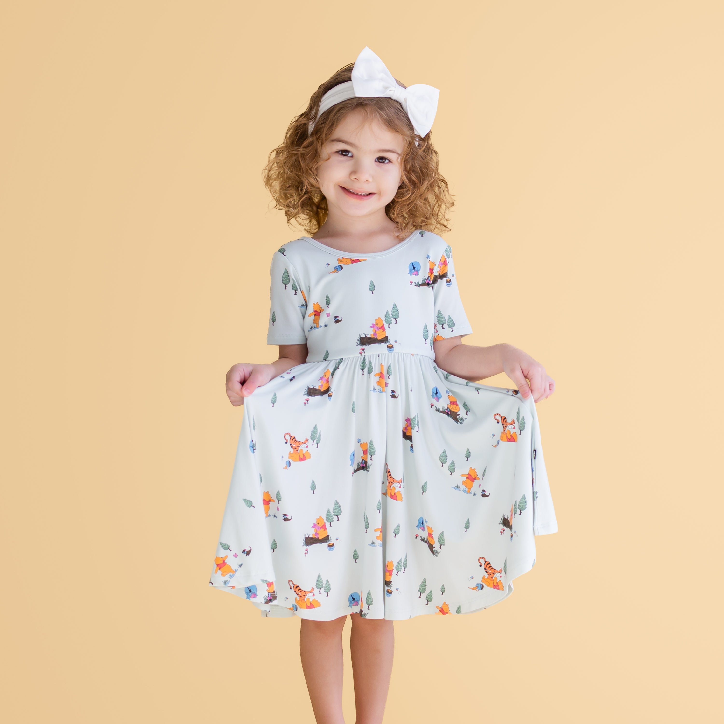 Young girl wearing the Twirl Dress in Winnie the Pooh Hundred Acre Wood holding the skirt of the dress out paired with a white bow