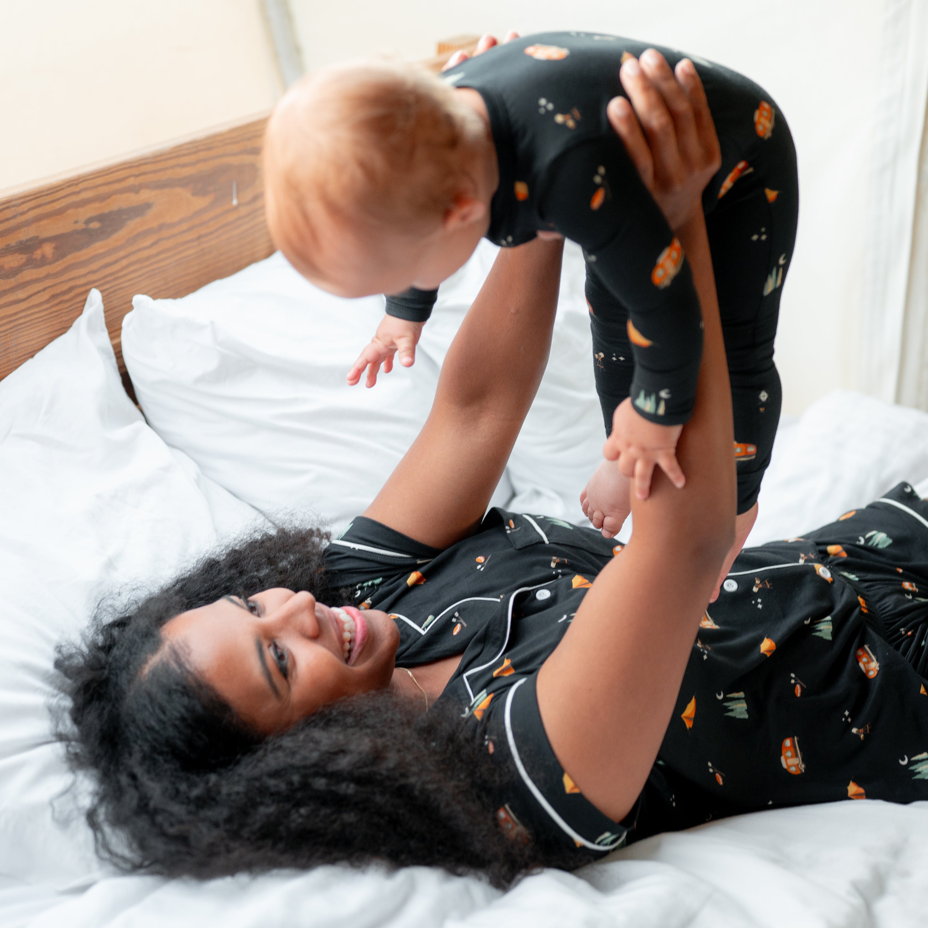 A mother laying in bed wearing the Women's Short Sleeve Pajama Set in Under the Stars holding up her child in the air who is in a matching romper