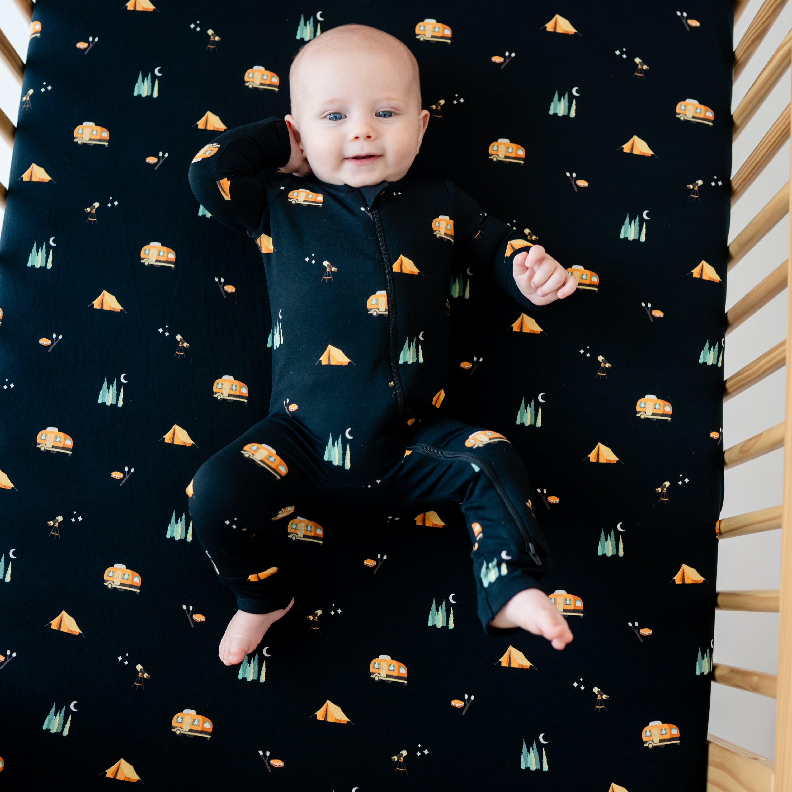 Infant laying in a crib wearing the Zippered Romper in Under the Stars laying on a matching crib sheet