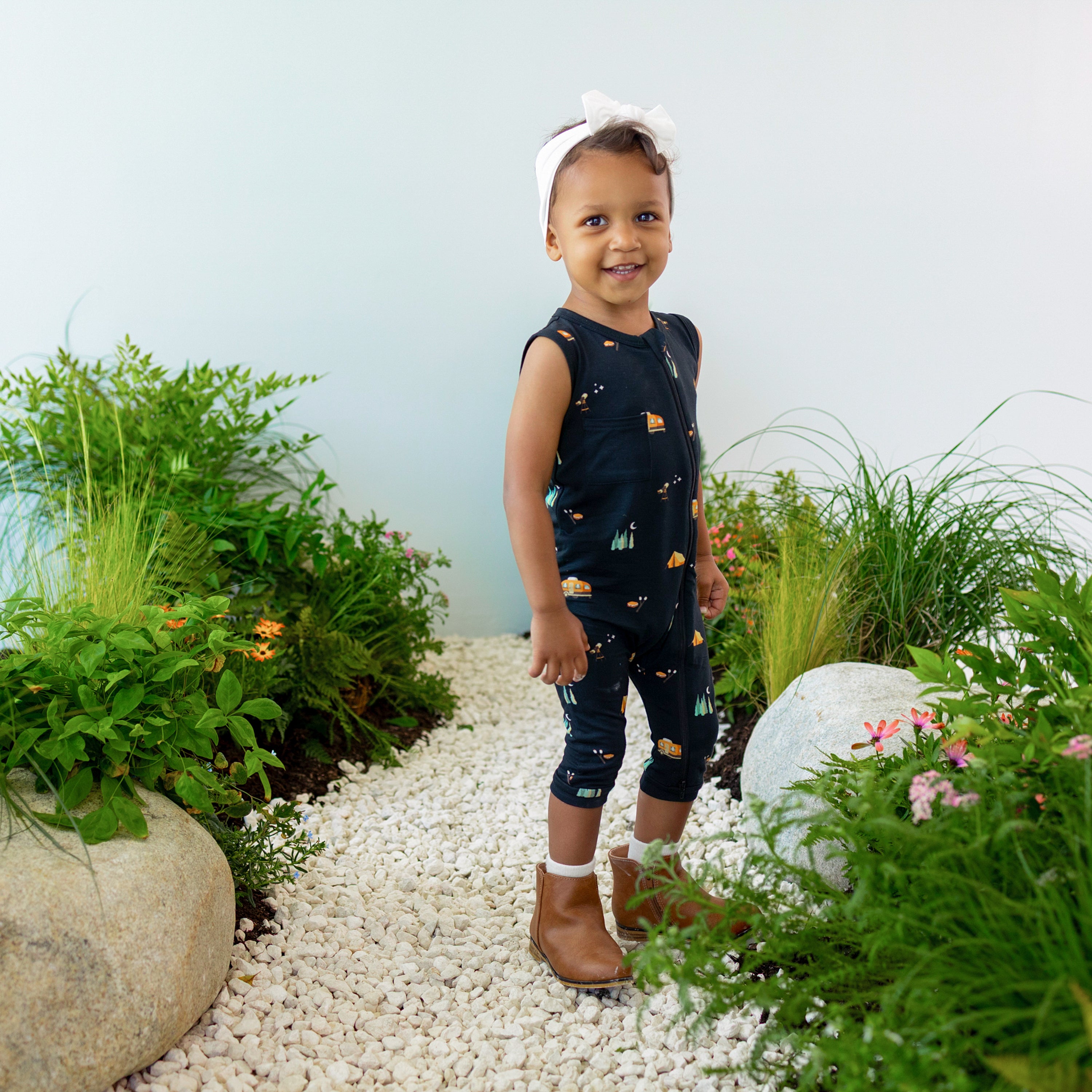 Smiling toddler wearing the Zippered Sleeveless Romper in Under the Stars with a Cloud Headband Bow standing on rocks surrounded by greenery
