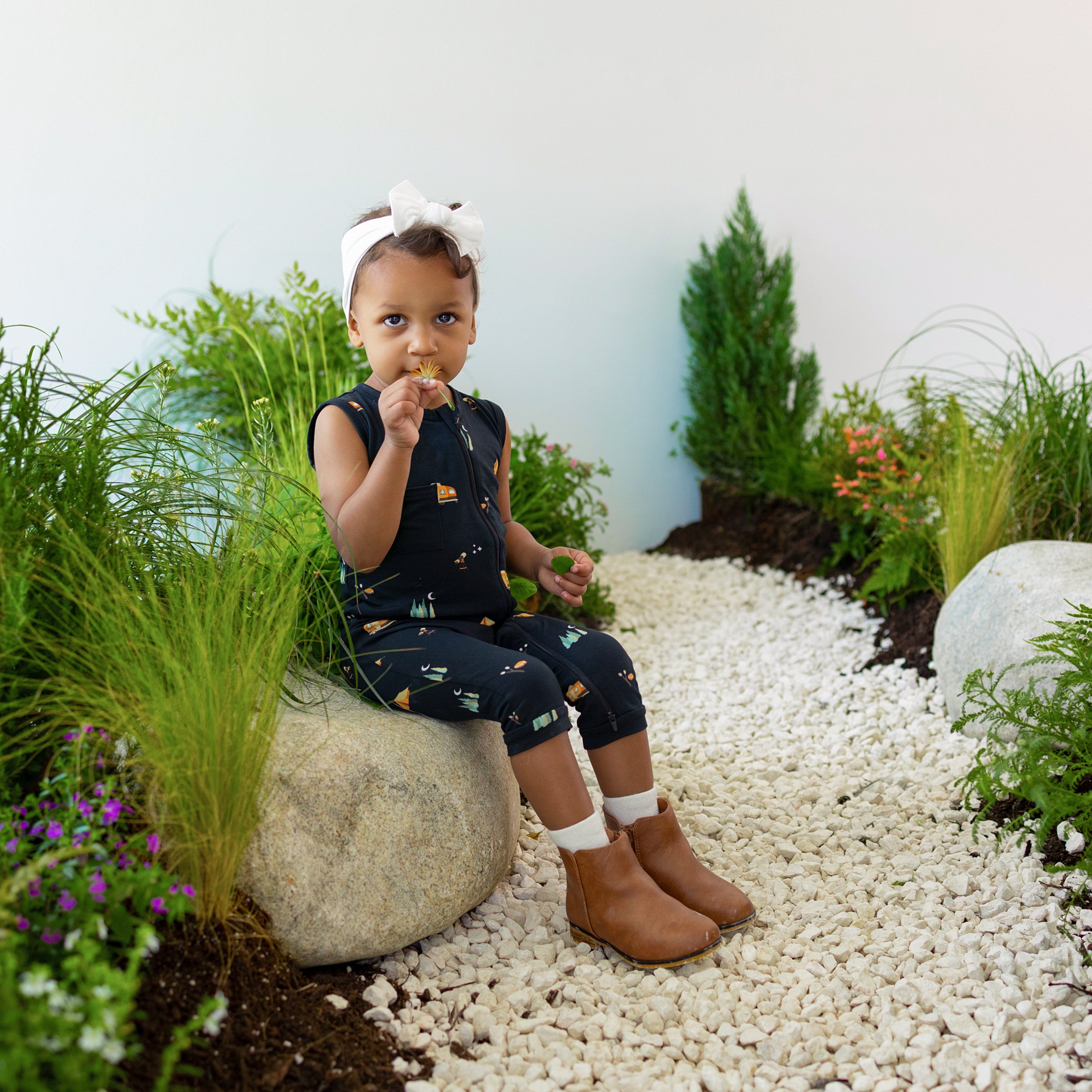 Young toddler sitting on a large rock smelling a flower wearing the Zippered Sleeveless Romper in Under the Stars