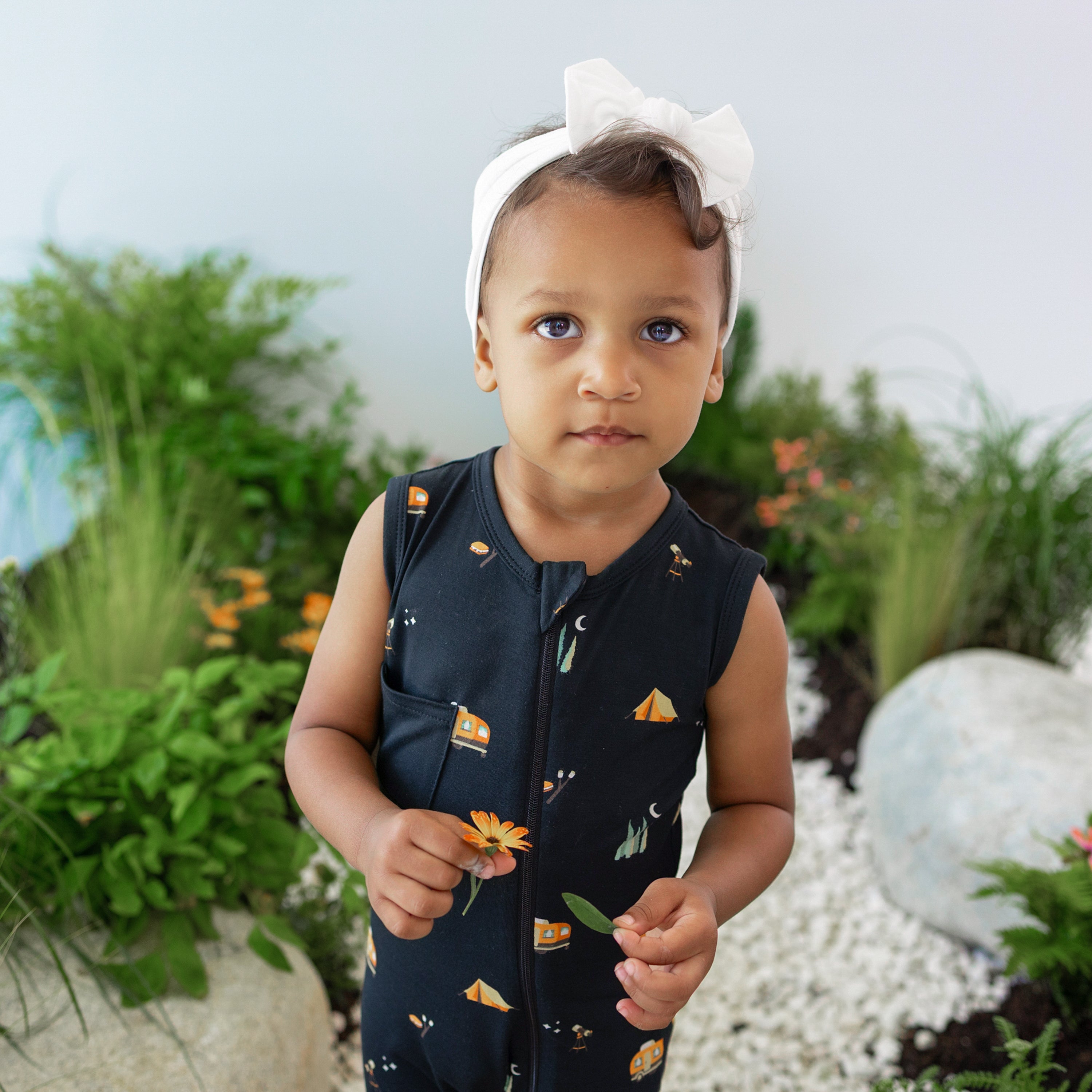 Close up of young toddler wearing the Zippered Sleeveless Romper in Under the Stars paired with a Cloud Headband Bow holding a leaf and a flower