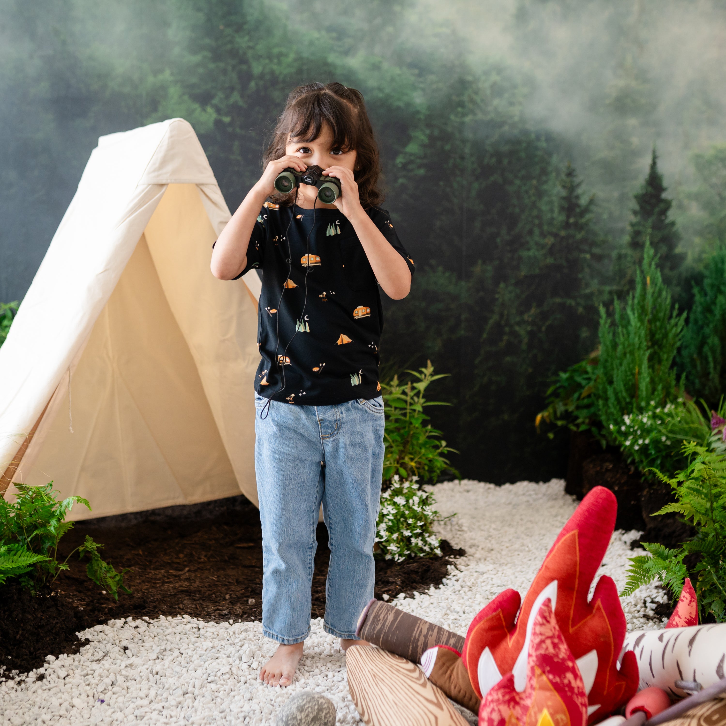 Young girl holding binoculars wearing the Zippered Shortall Romper in Under the Stars paired with jeans standing in rock in front of a campsite backdrop