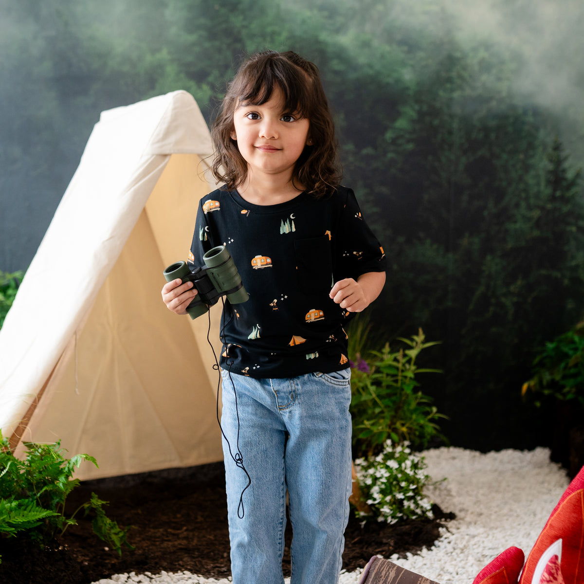 Girl toddler wearing the Zippered Shortall Romper in Under the Stars paired with jeans while holding binoculars and standing in front of a wilderness campsite back drop