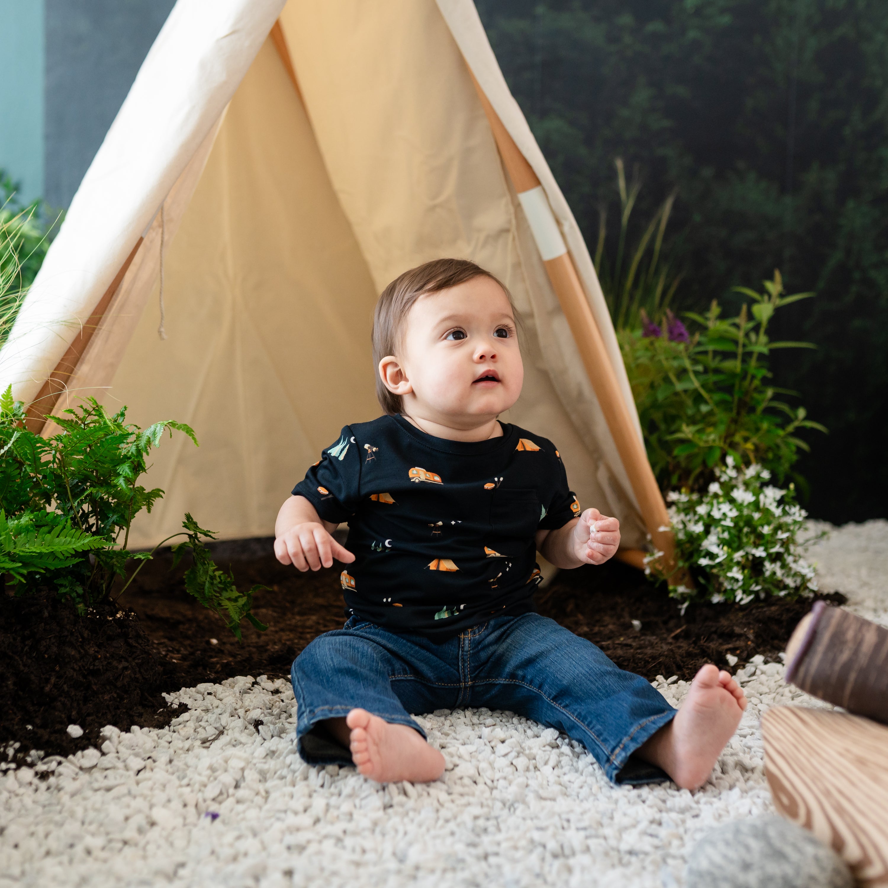 Infant modeling the Toddler Crew Neck Tee in Under the Stars paired with jeans while sitting in rocks with a camping backdrop