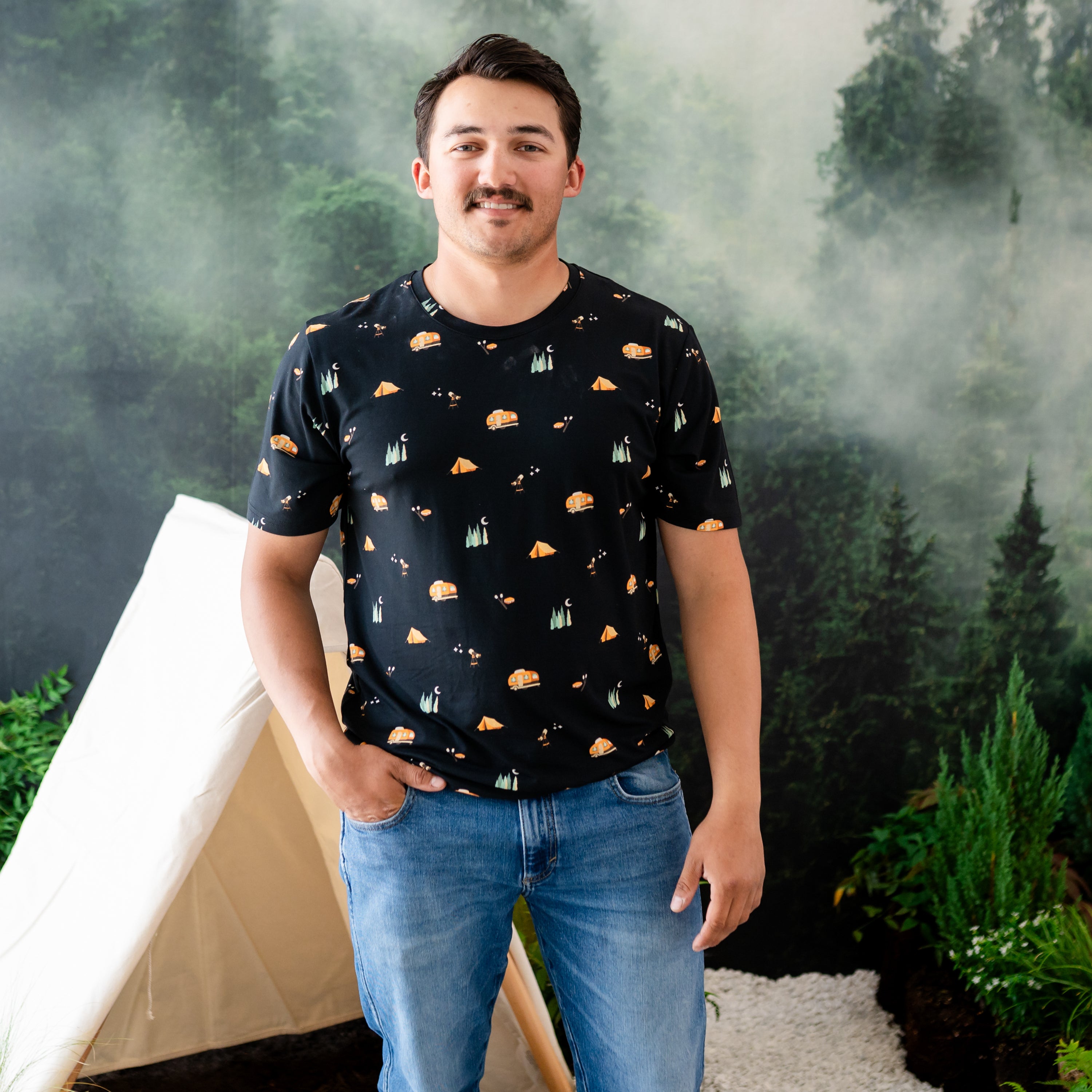 Male model standing in front of a camping backdrop wearing the Men's Crew Neck Tee in Under the Stars paired with jeans with one hand in his pocket