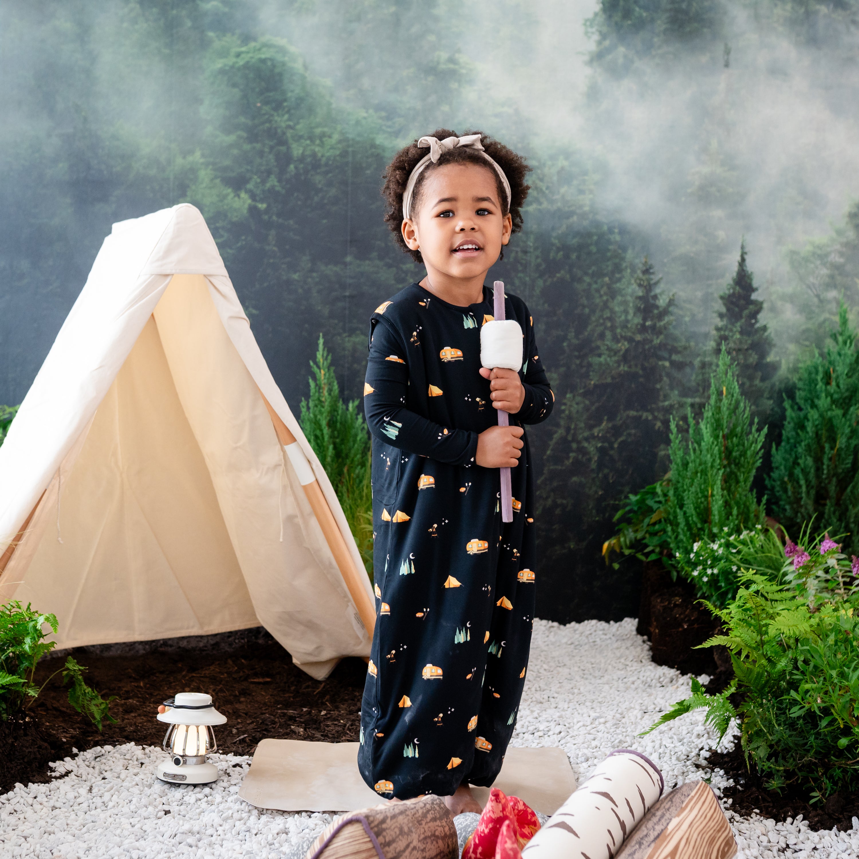 Child standing wearing the Sleep Bag Walker in Under the Stars 1.0 holding a toy stick with a marshmellow on it surrounded by a wilderness inspired camping back drop