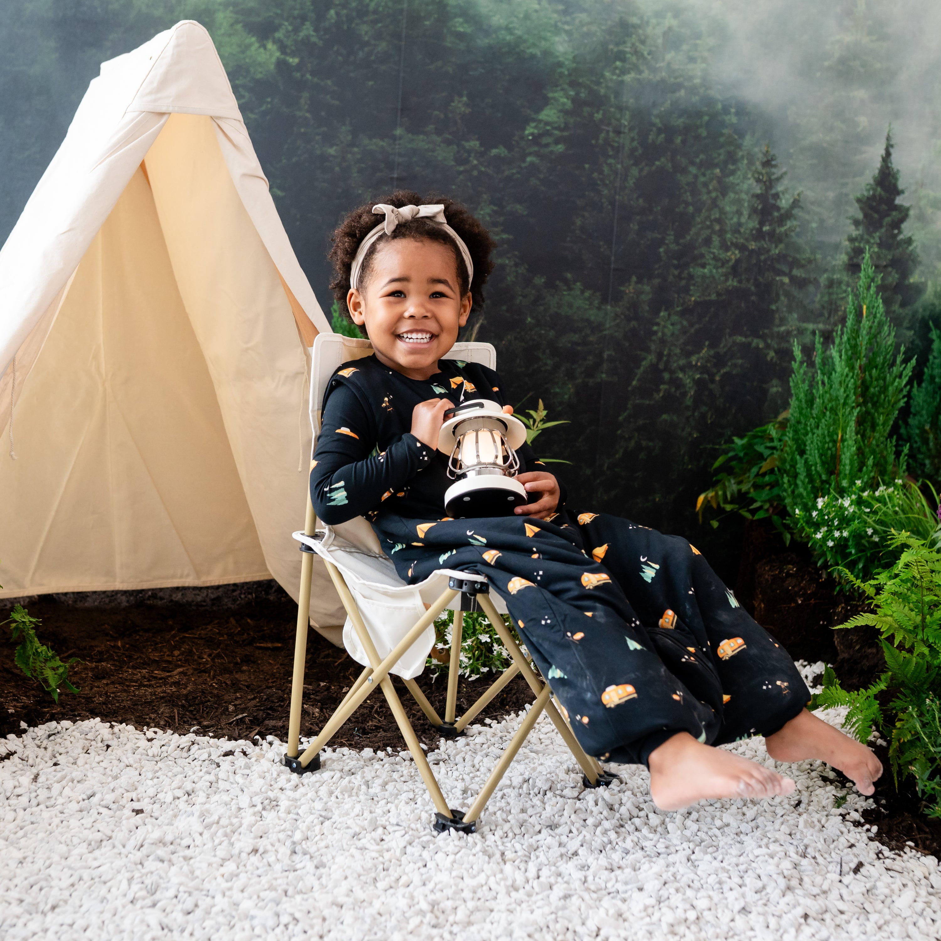 Child model sitting in a canvas camping chair holding a light lantern wearing the Sleep Bag Walker in Under the Stars 1.0