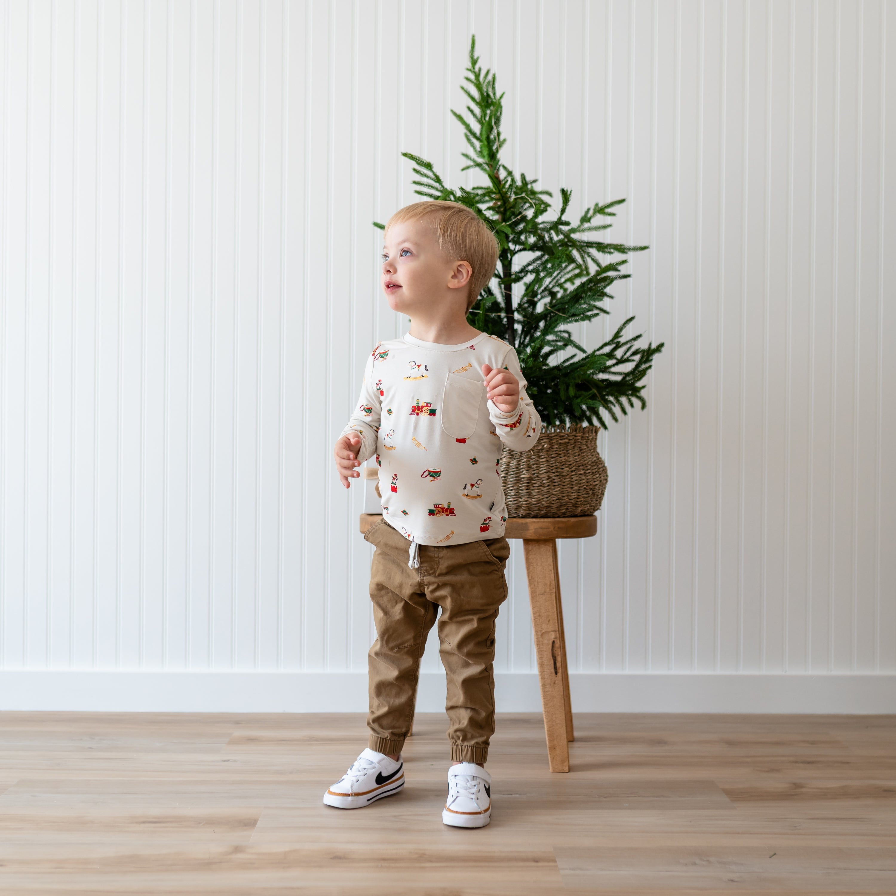 Young boy standing in front of a potted plant in a wicker basket on a wood stool wearing the Long Sleeve Toddler Crew Neck Tee in Vintage Toys with dark tan pants