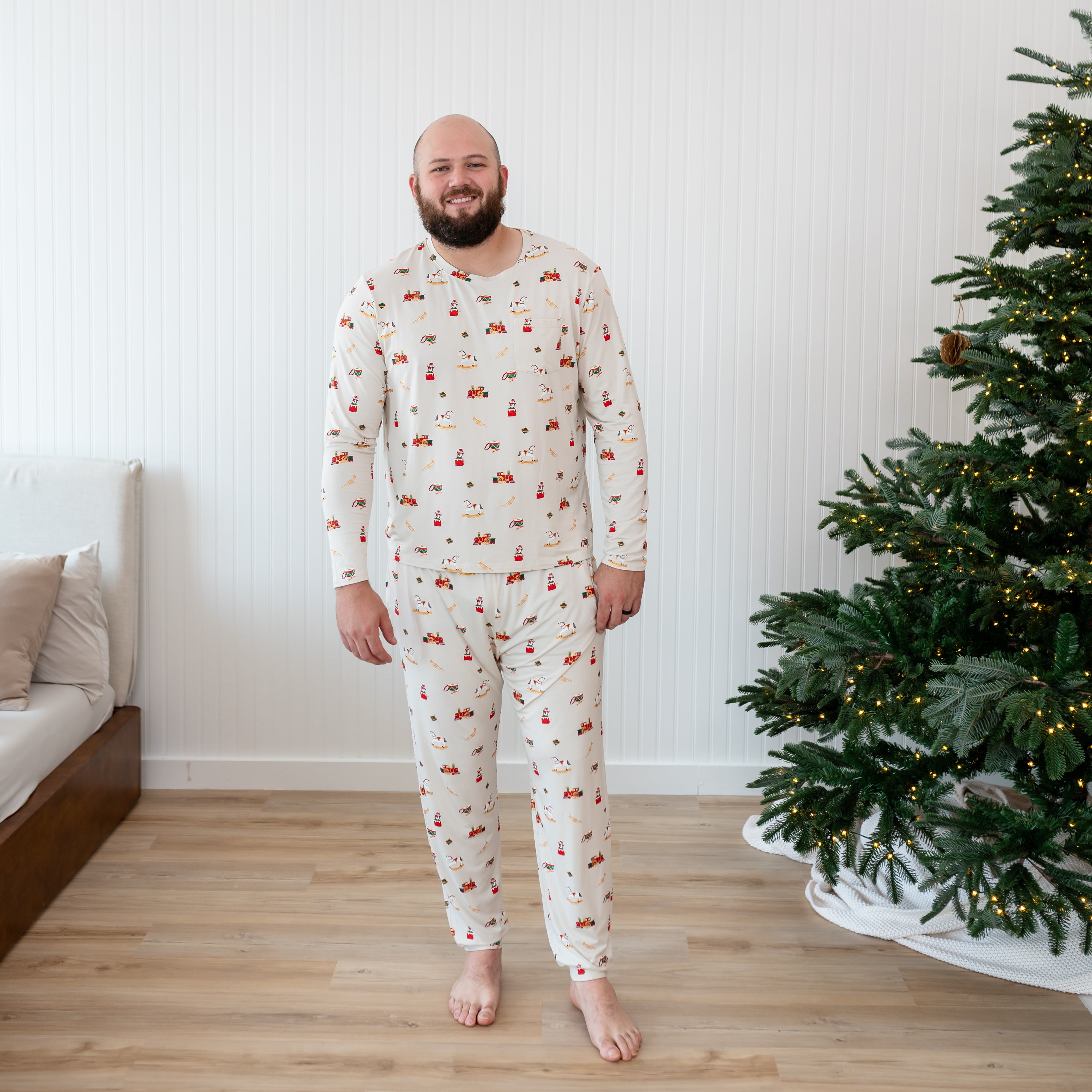Smiling male model standing beside a lit Christmas Tree in front of a paneled wall wearing the Men's Jogger Set in Vintage Toys