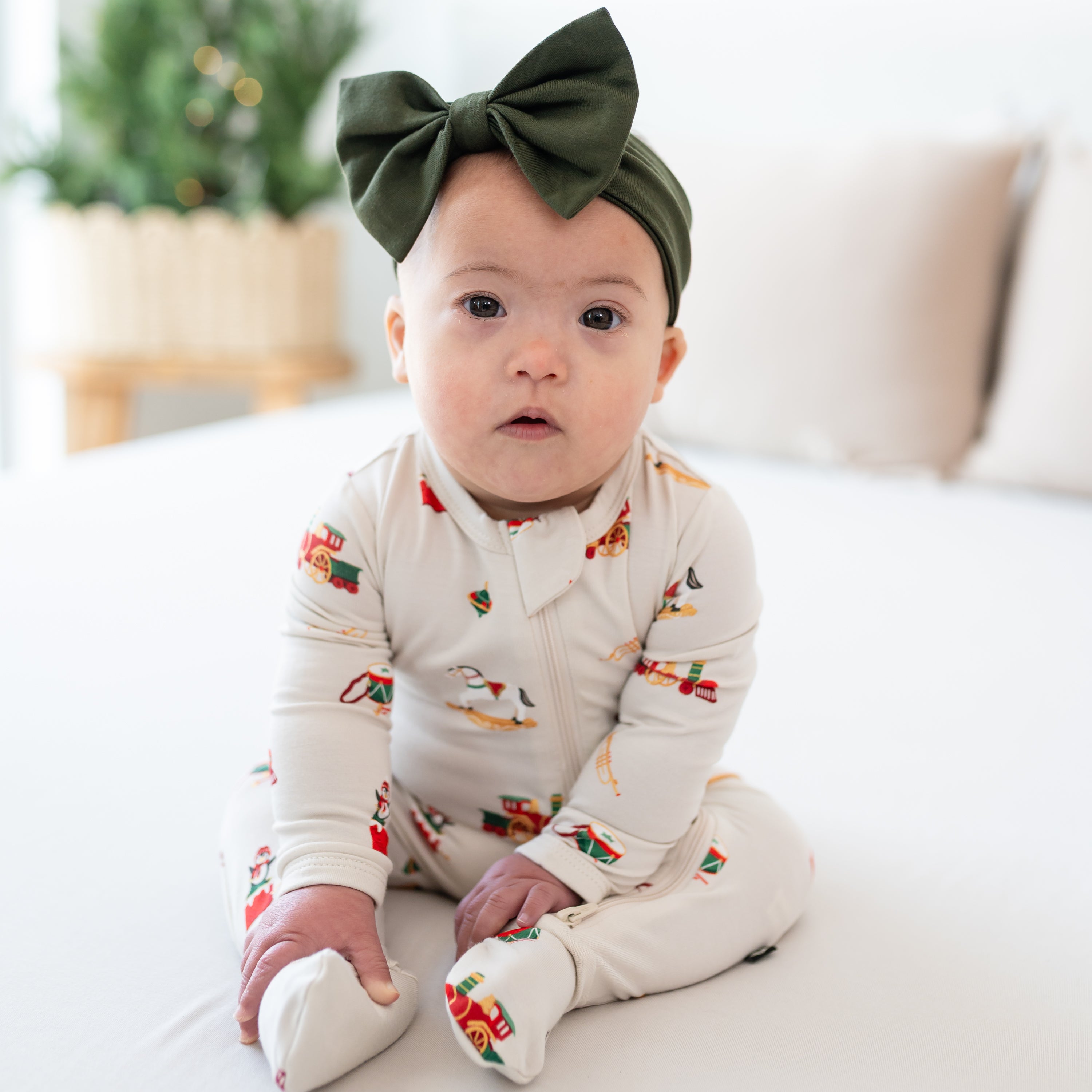 Young infant girl sitting on a bed wearing the Zippered Footie in Vintage Toys paired with a Fir Bow Headband