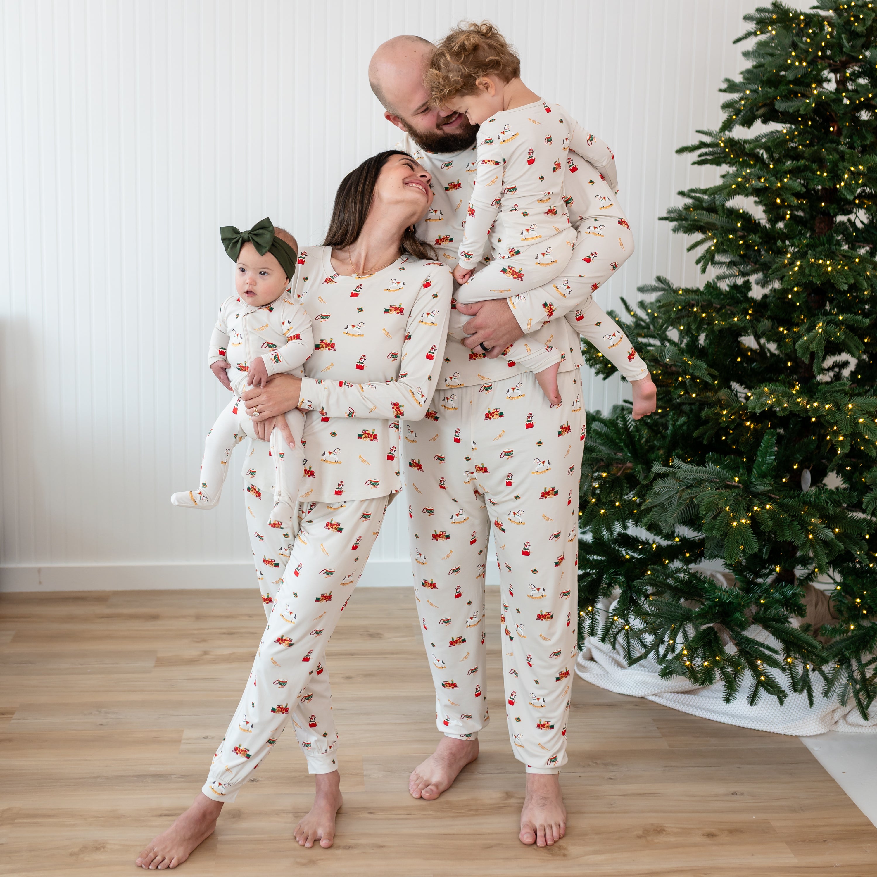Family of 4 matching in Vintage Toys Pajamas standing beside a lit Christmas tree