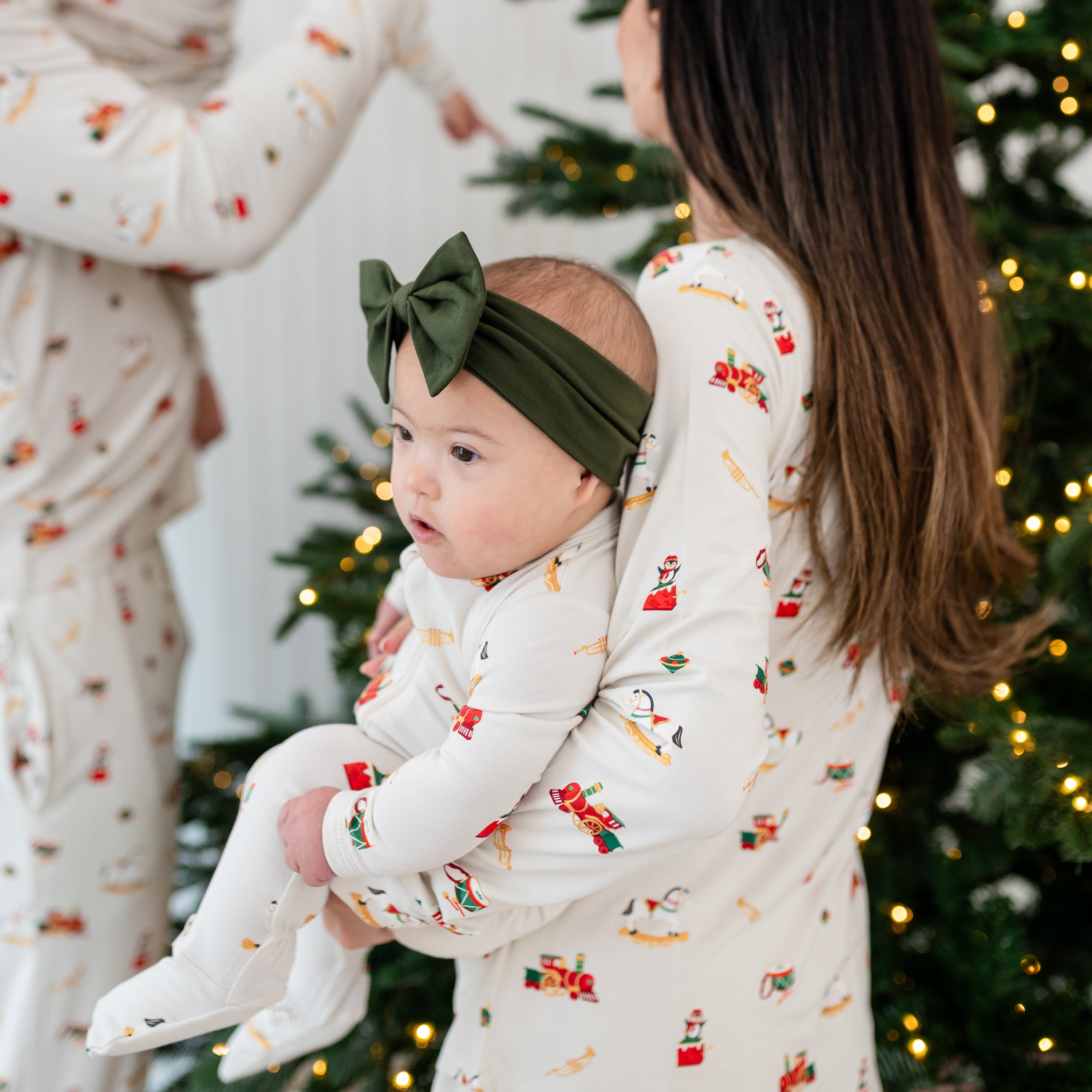 Young infant wearing the Zippered Footie in Vintage Toys and Fir Bow Headband being held by her mother matching in the Women's Jogger Pajama Set in Vintage Toys