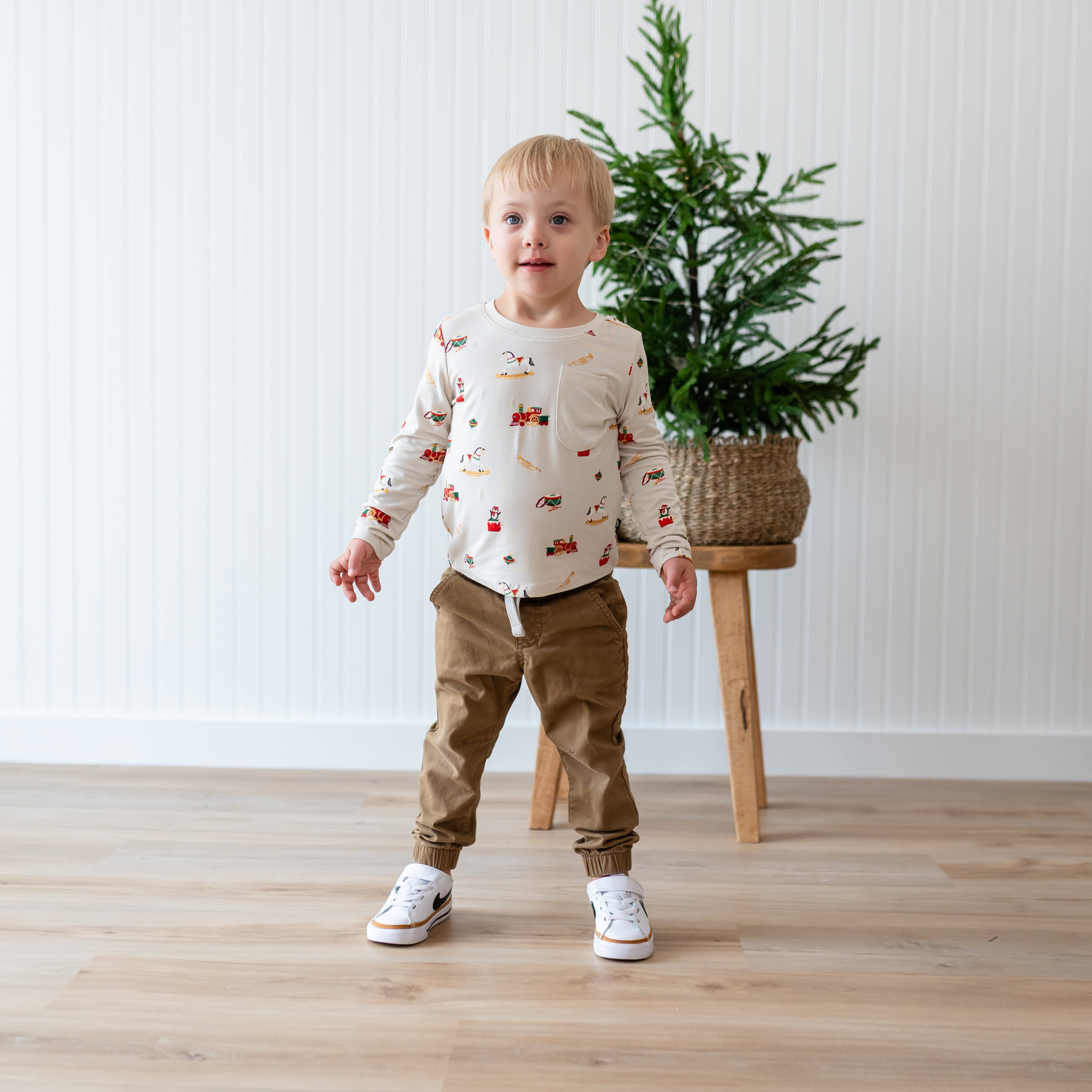 Young boy wearing the Long Sleeve Toddler Crew Neck Tee in Vintage Toys standing in front of a potted plant resting on a wooden stool