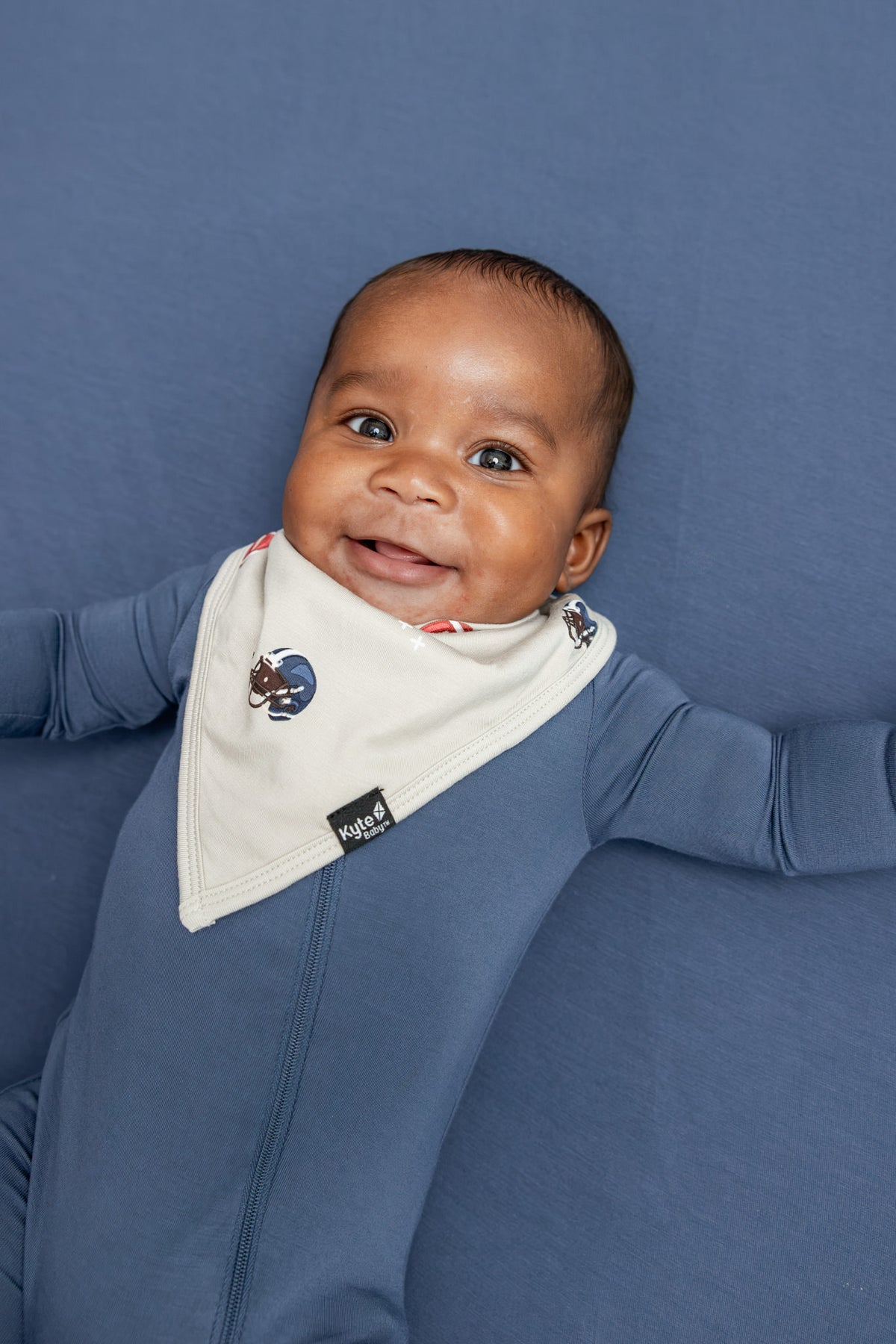 A smiling baby wearing a blue long-sleeve footie and a white bib with small football graphics, lying on a blue textured background.