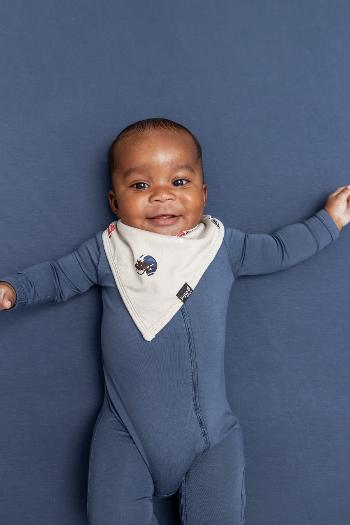 A baby wearing a slate blue footie and a cream bib with a football pattern, lying on a solid slate blue background