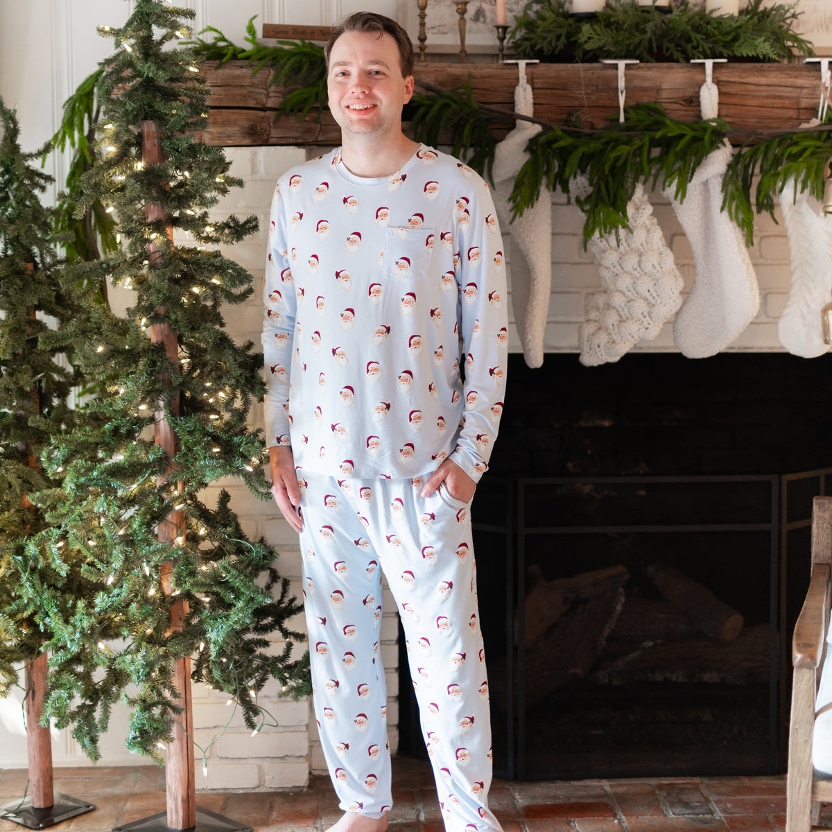 Smiling male model standing in front of a white brick fireplace with white stockings hung on the mantel wearing the Men's Jogger Set in Vintage Santa with one hand in the pants pocket
