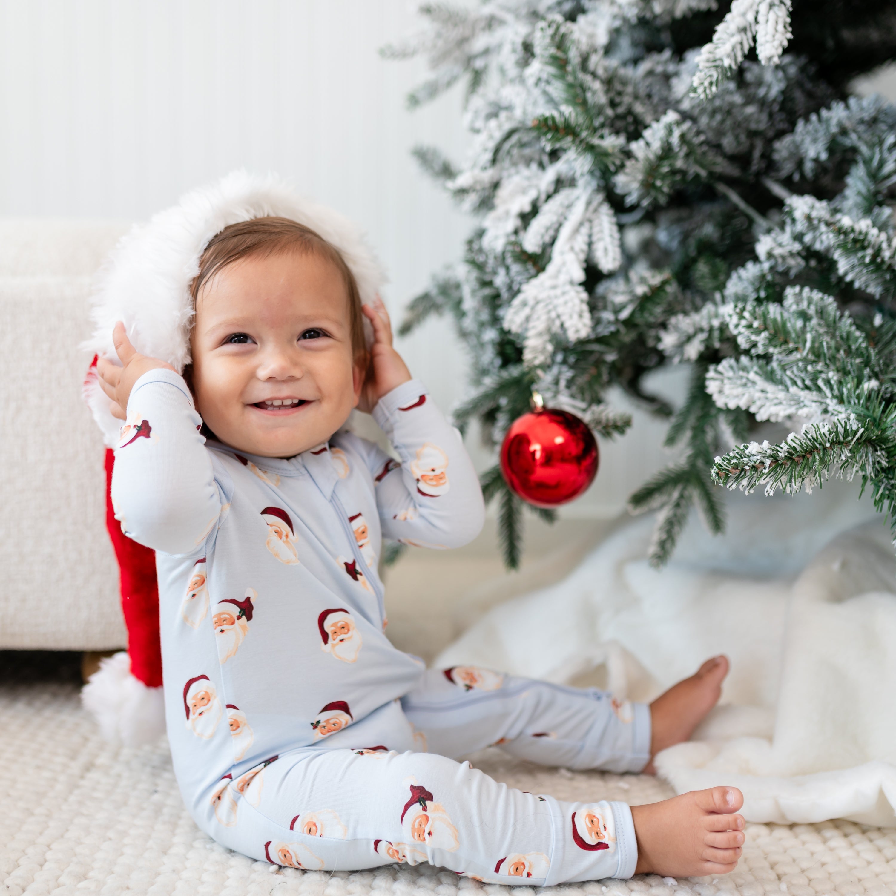 Young smiling toddler sitting beside a frosted Christmas tree wearing the Zippered Romper in Vintage Santa with a Santa hat