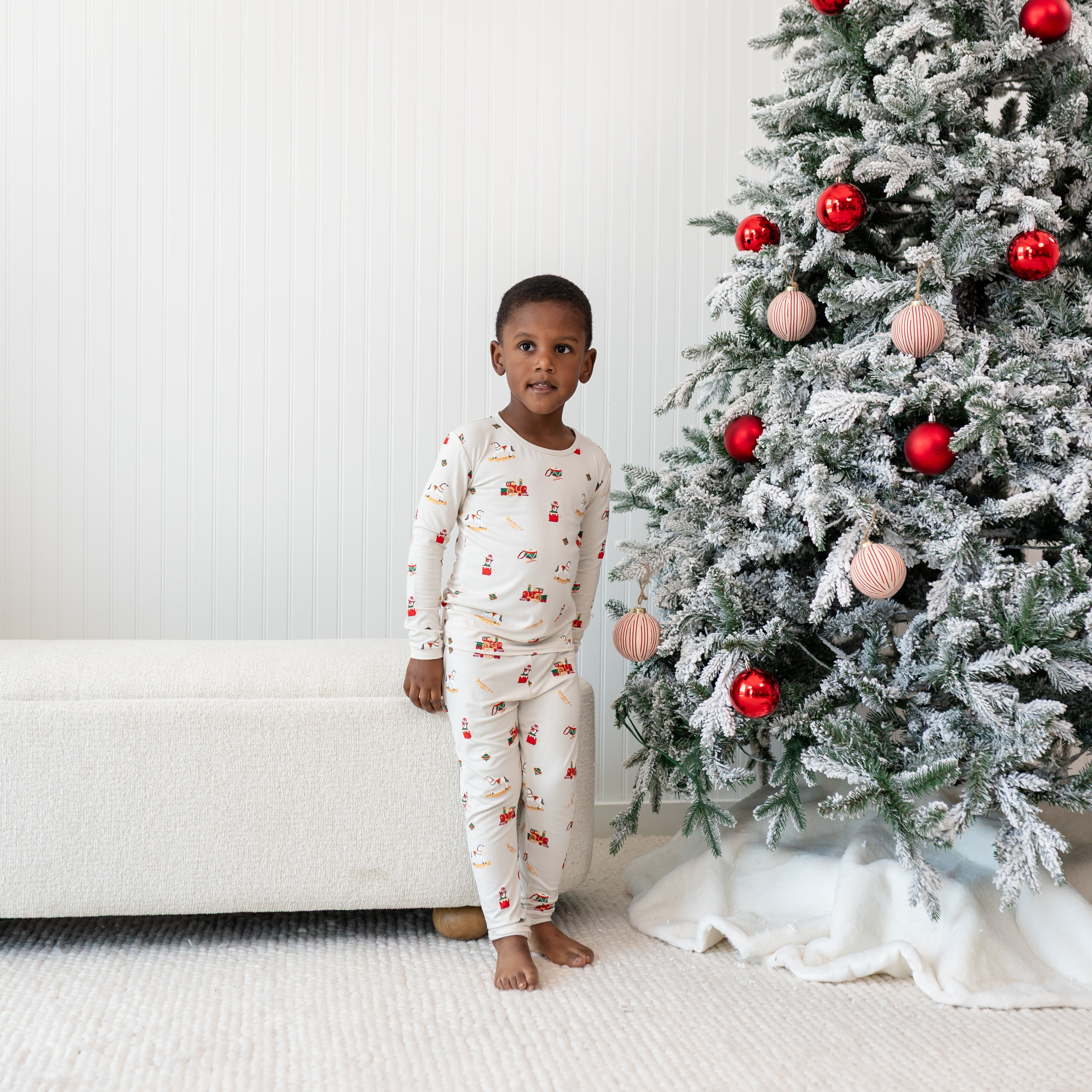 Young boy standing in front of a cloth bench beside a decorated Christmas Tree wearing the Long Sleeve Pajamas in Vintage Toys
