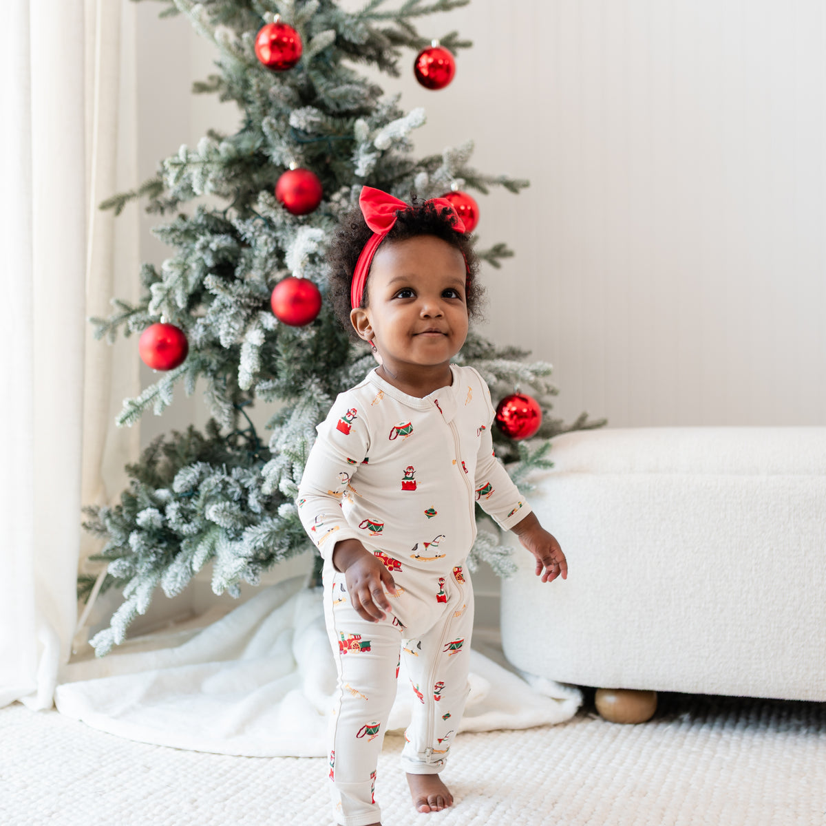 Young toddler standing in front of a decorated Christmas tree wearing the Zippered Romper in Vintage Toys with a red bow headband