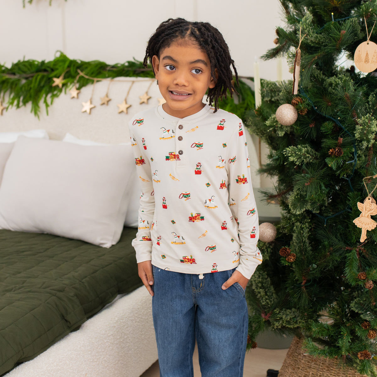 Young girl standing between a decorated Christmas tree and bed wearing the Long Sleeve Toddler Henley Tee in Vintage Toys with one hand in her jeans pocket