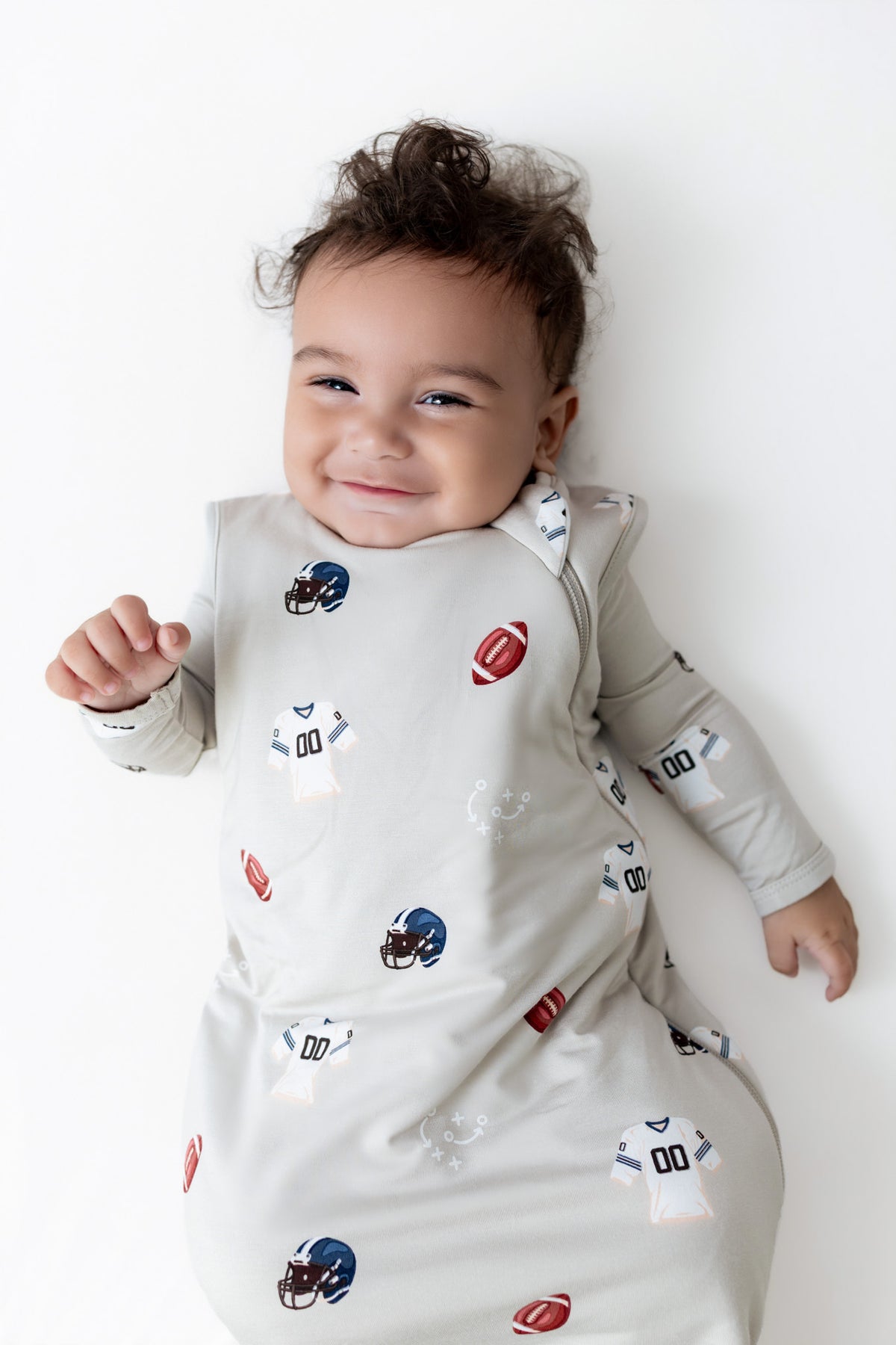 A baby lying on their back wearing a light gray long-sleeve sleep sack with a pattern of football helmets and footballs, against a plain white background.