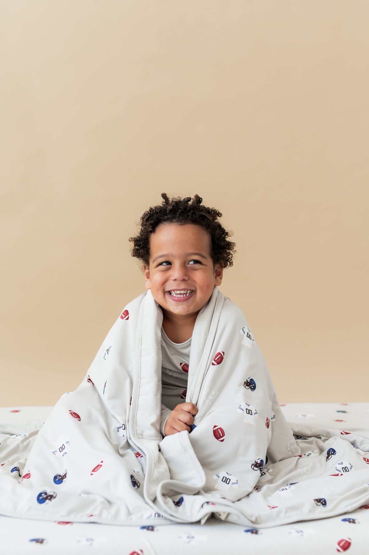 Boy holding Toddler Blanket in Vintage Football 1.0 print covered over his shoulders sitting on a similar print sheet on a beige background
