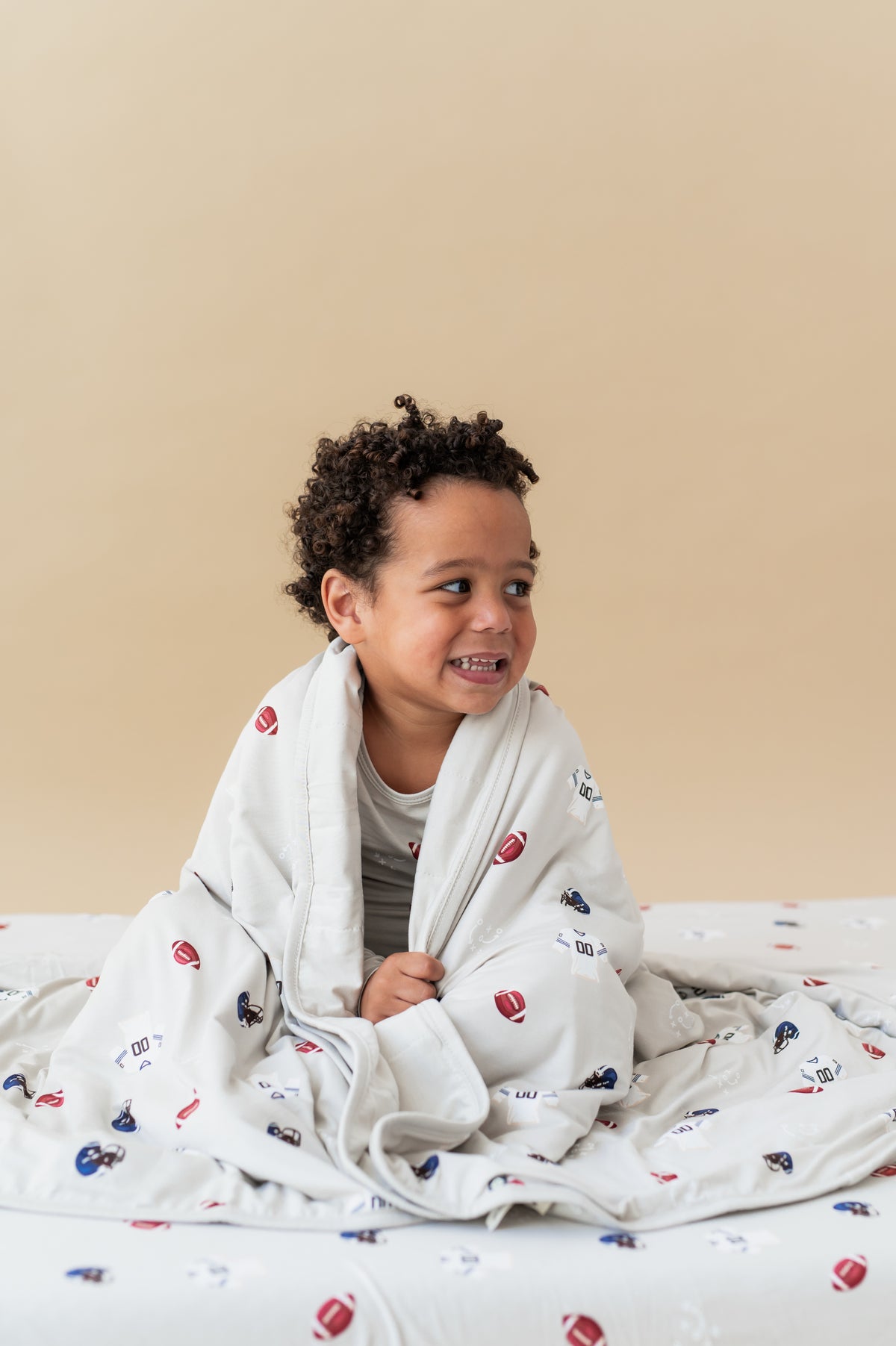 Boy holding Toddler Blanket in Vintage Football 1.0 print covered over his shoulders sitting on a similar print sheet on a beige background