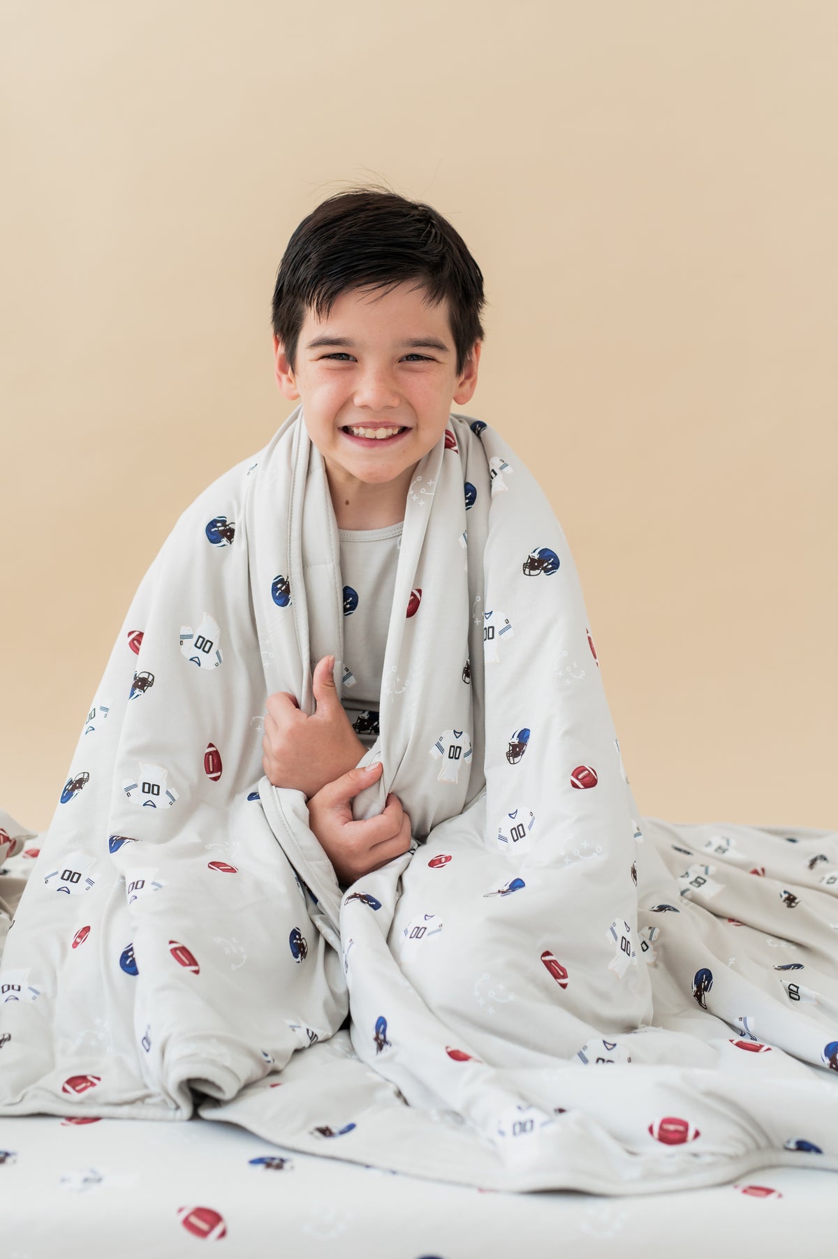 Boy holding Youth Blanket in Vintage Football 2.5 print over his shoulders sitting on a similar print sheet on a beige background