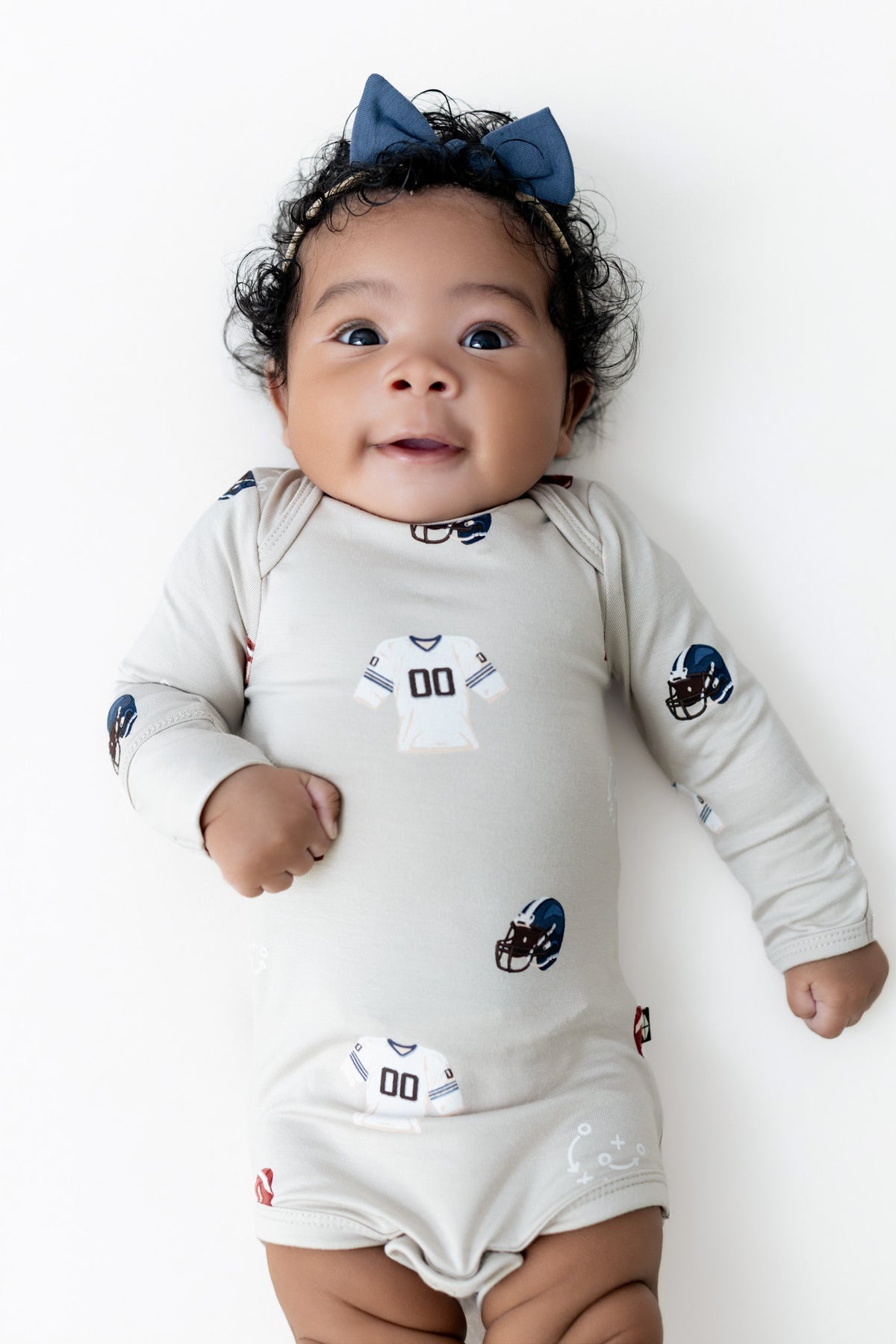 A baby lying on a white background wearing a long-sleeve cream bodysuit with NFL football jersey graphics and a blue bow headband.