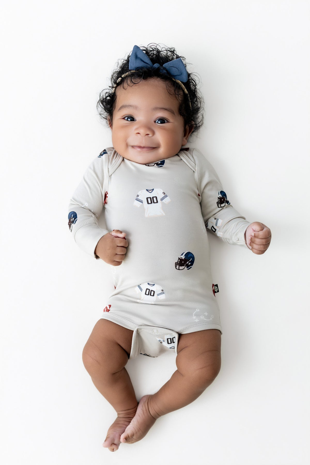 A baby lying on a white background wearing a long-sleeve cream-colored romper with small football-themed prints and a matching blue bow headband.