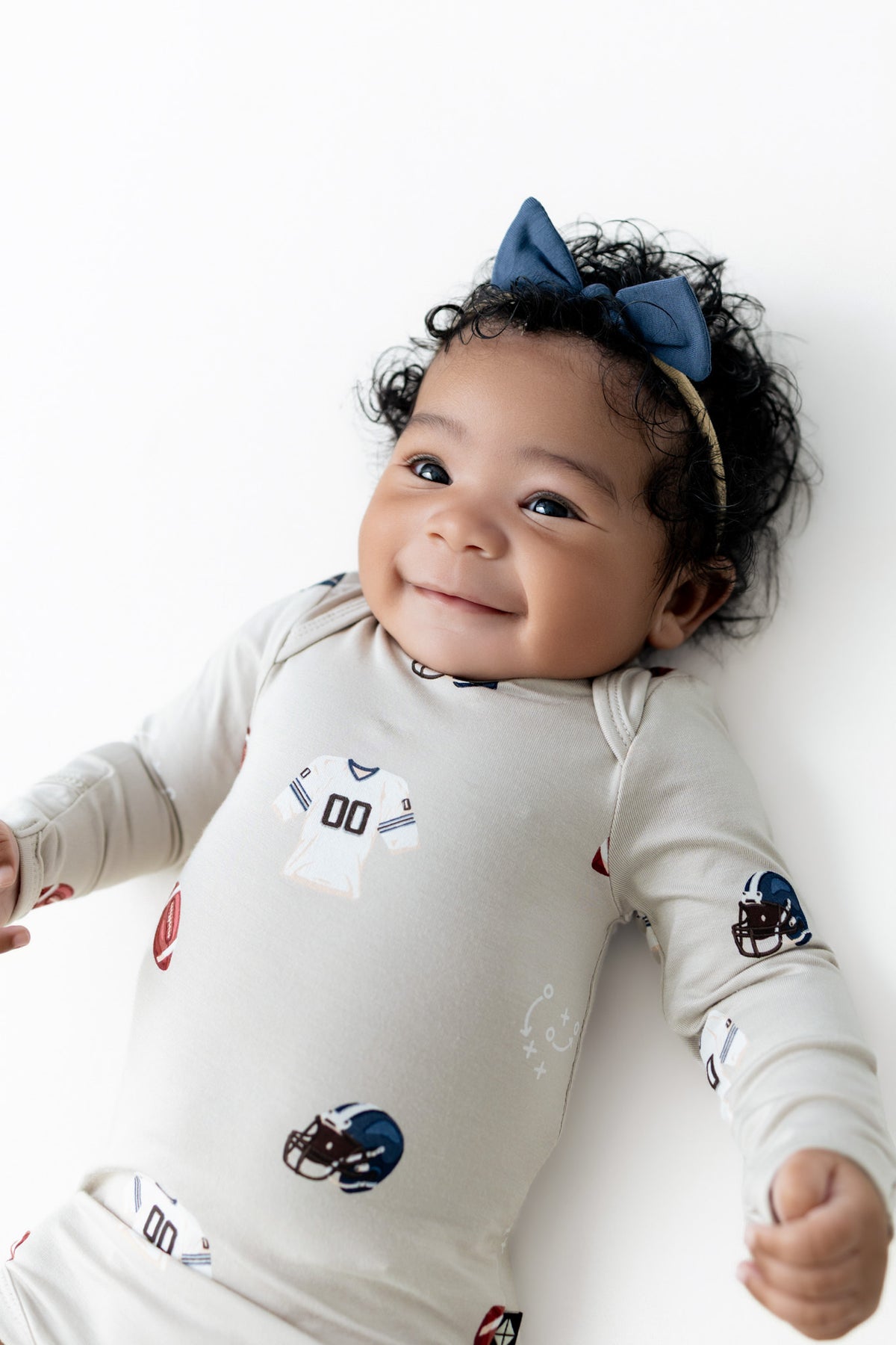 A smiling baby lying on their back wearing a cream long-sleeve bodysuit with football jersey and helmet prints, along with a blue bow headband, against a clean white background.