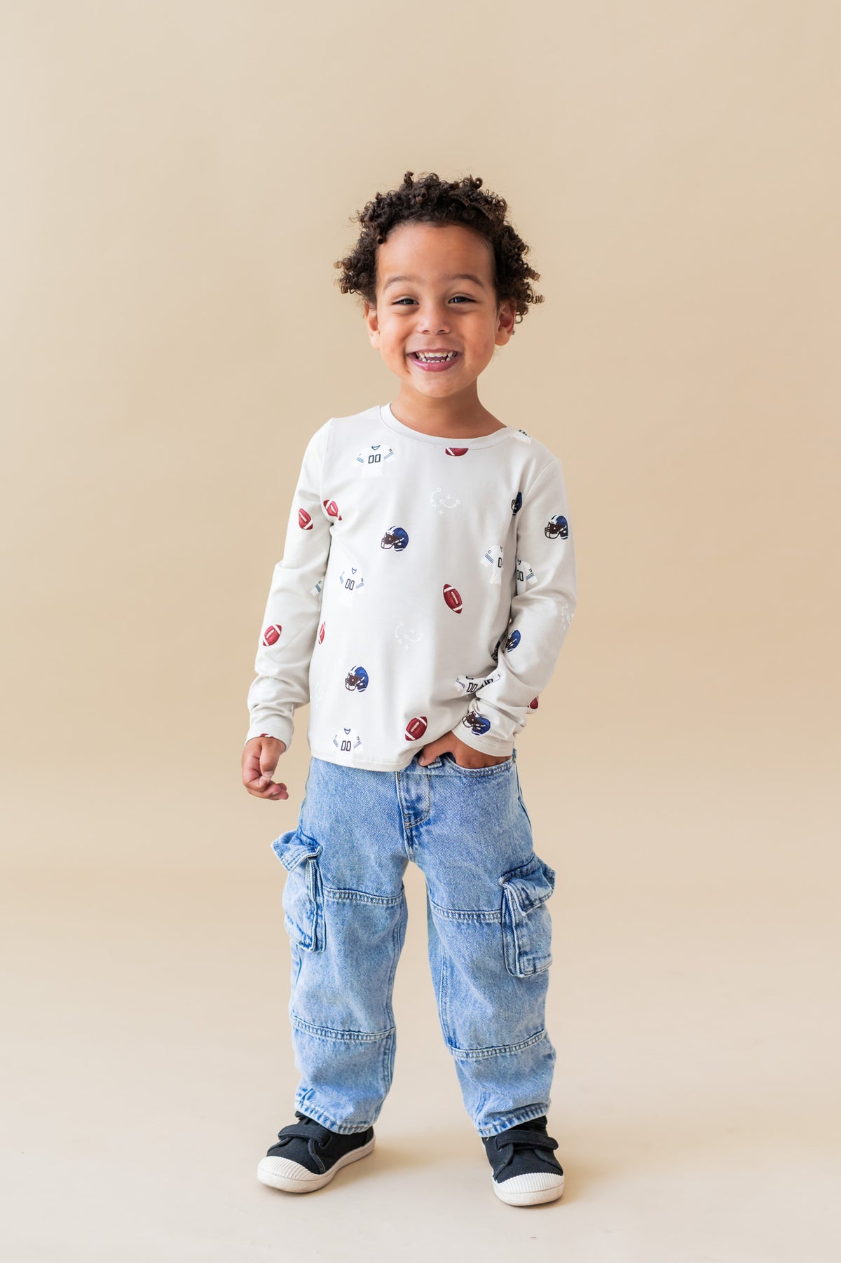 Boy posing in Long Sleeve Toddler Crew Neck Tee in Vintage Football print over blue jeans on a beige background