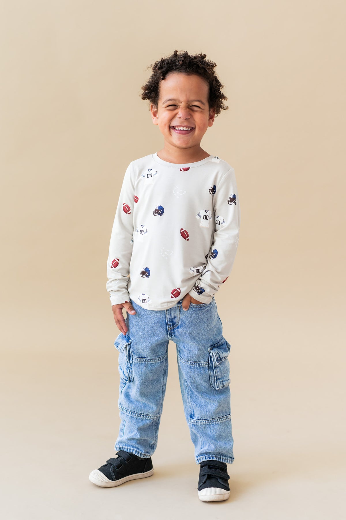 Boy smiling in Long Sleeve Toddler Crew Neck Tee in Vintage Football print over blue jeans on a beige background