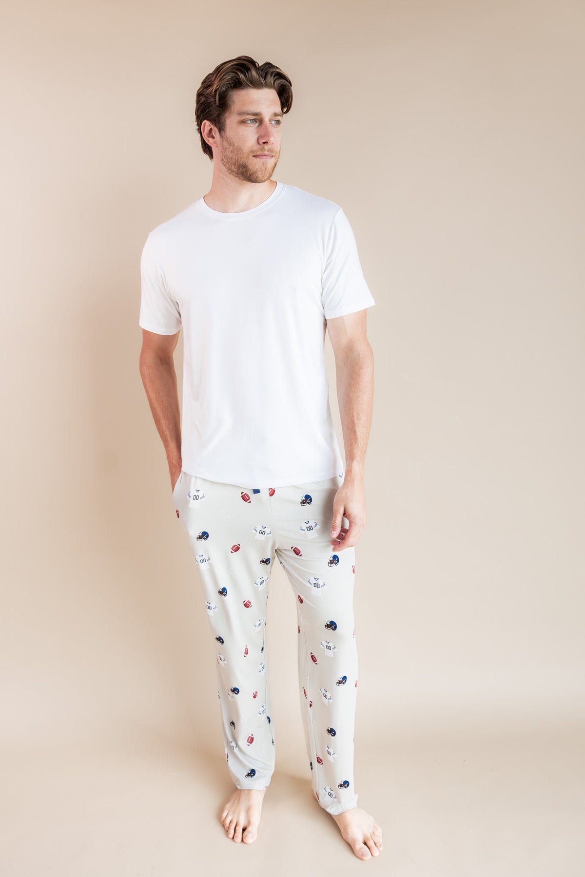 A man modeling a white crewneck t-shirt and white pajama pants with a small red, white, and blue football pattern, standing against a solid beige studio background.