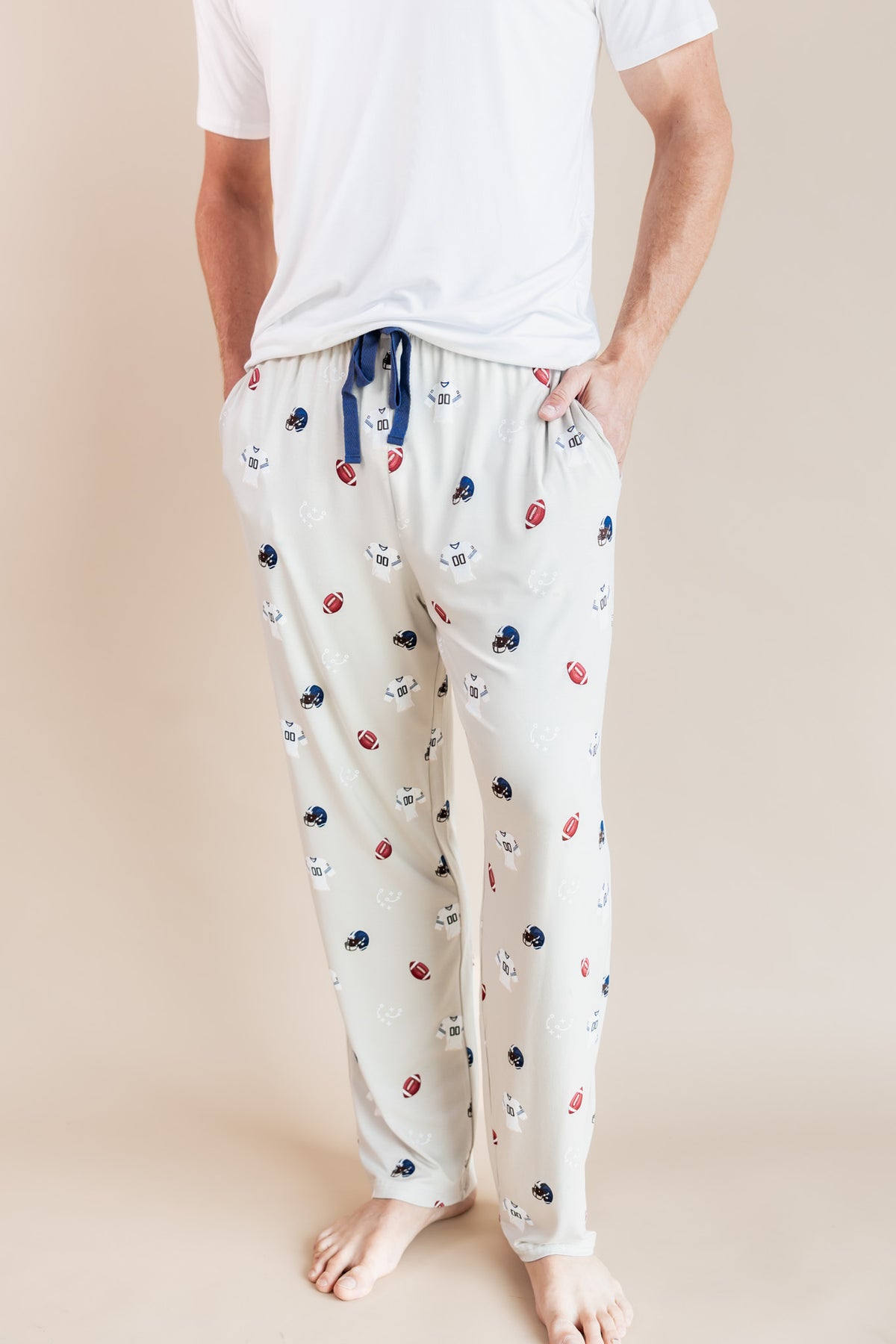 A person wearing cream-colored pajama pants with a small multicolor pattern of footballs and helmets, paired with a white t-shirt, standing against a solid beige background.