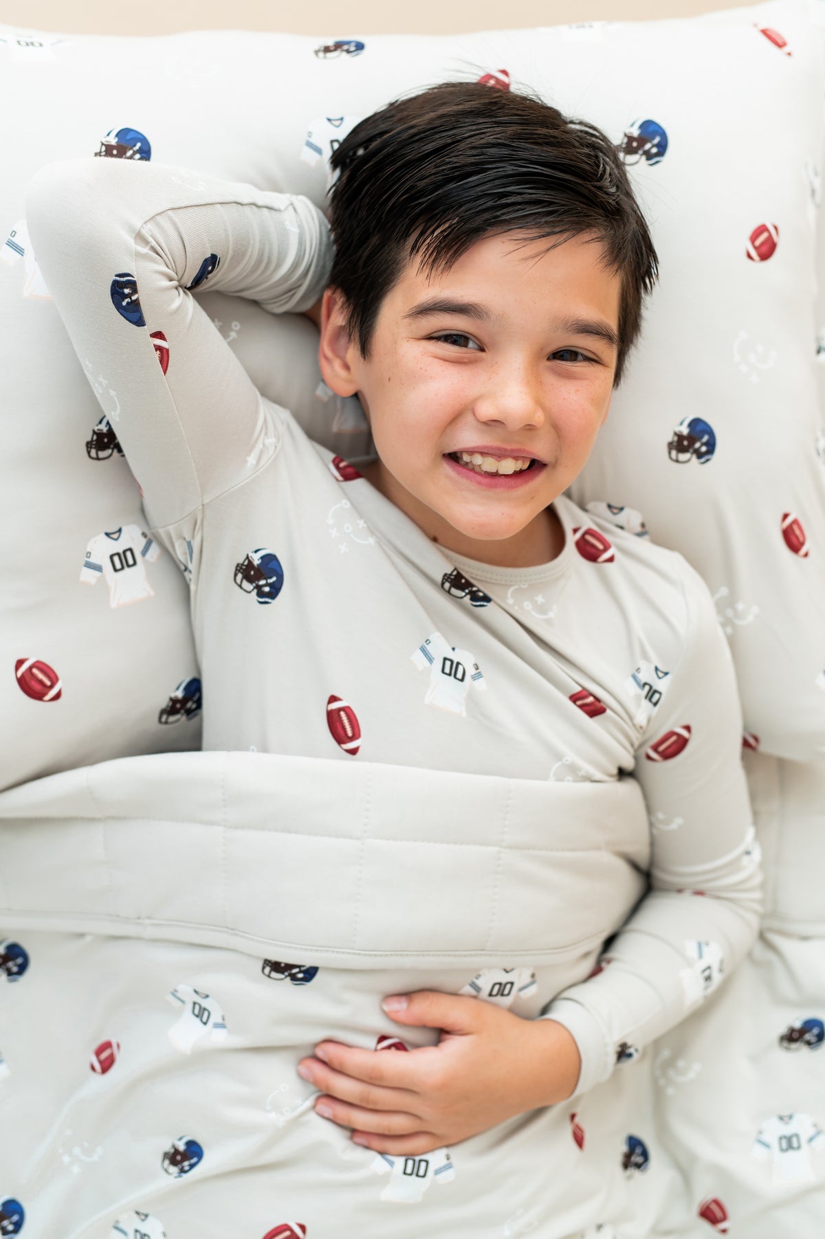 Boy lying on Standard Pillowcase and sheets in Vintage Football print, wearing a similar print Long sleeve pajama set covered under a similar print youth blanket