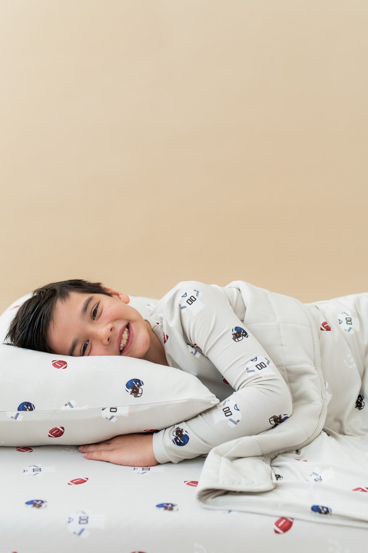 Boy lying on Standard Pillowcase and sheets in Vintage Football print, wearing a similar print Long sleeve pajama set covered similar print youth blanket on a beige background