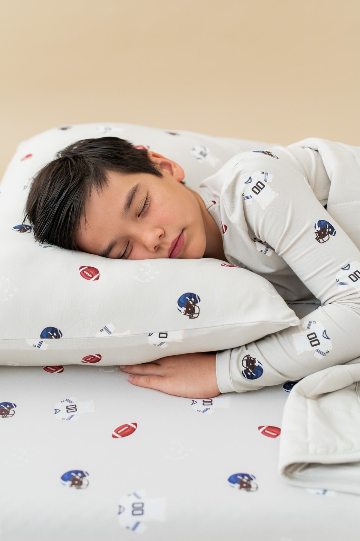 Boy sleeping on Standard Pillowcase and sheets in Vintage Football print wearing a similar print Long sleeve pajama set on a beige background