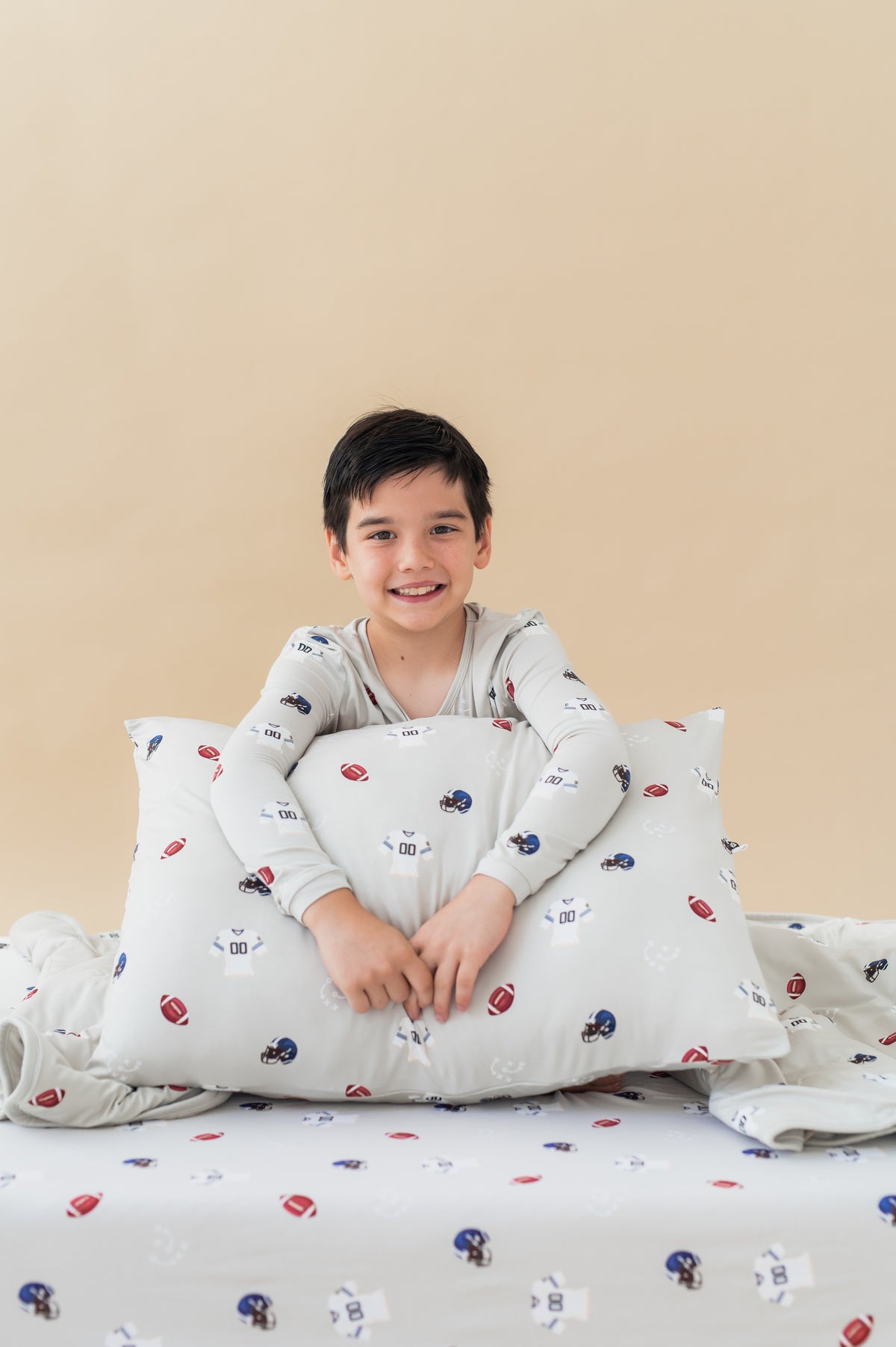 Boy holding Standard Pillowcase in Vintage Football print sitting on a similar print sheets, wearing a similar print Long sleeve pajama set on a beige background